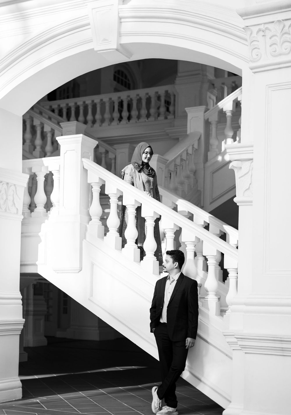 Black and white photo of a couple on an elegant staircase, man in suit below, woman in hijab above.