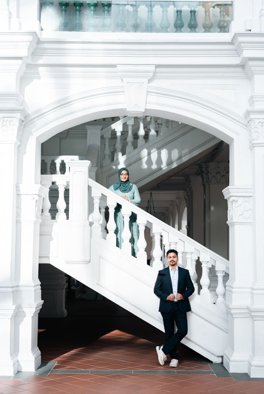 Couple posing on white ornate staircase in elegant colonial building, woman in hijab above, man in suit below.