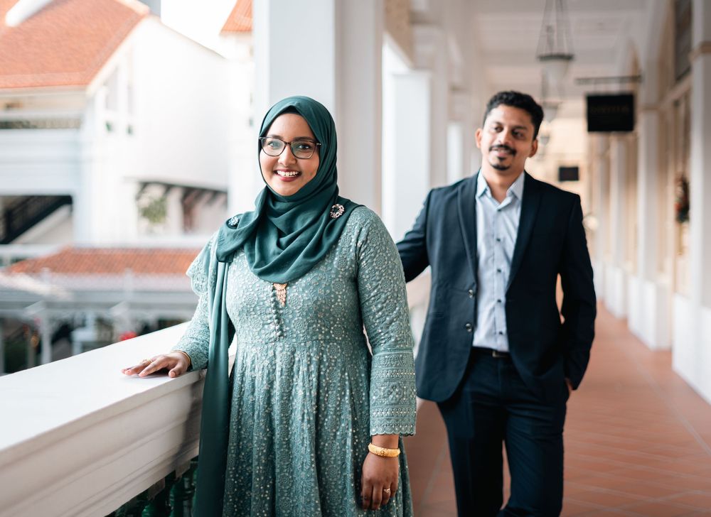 A woman in a green hijab and dress stands beside a man in a suit on a sunlit corridor.