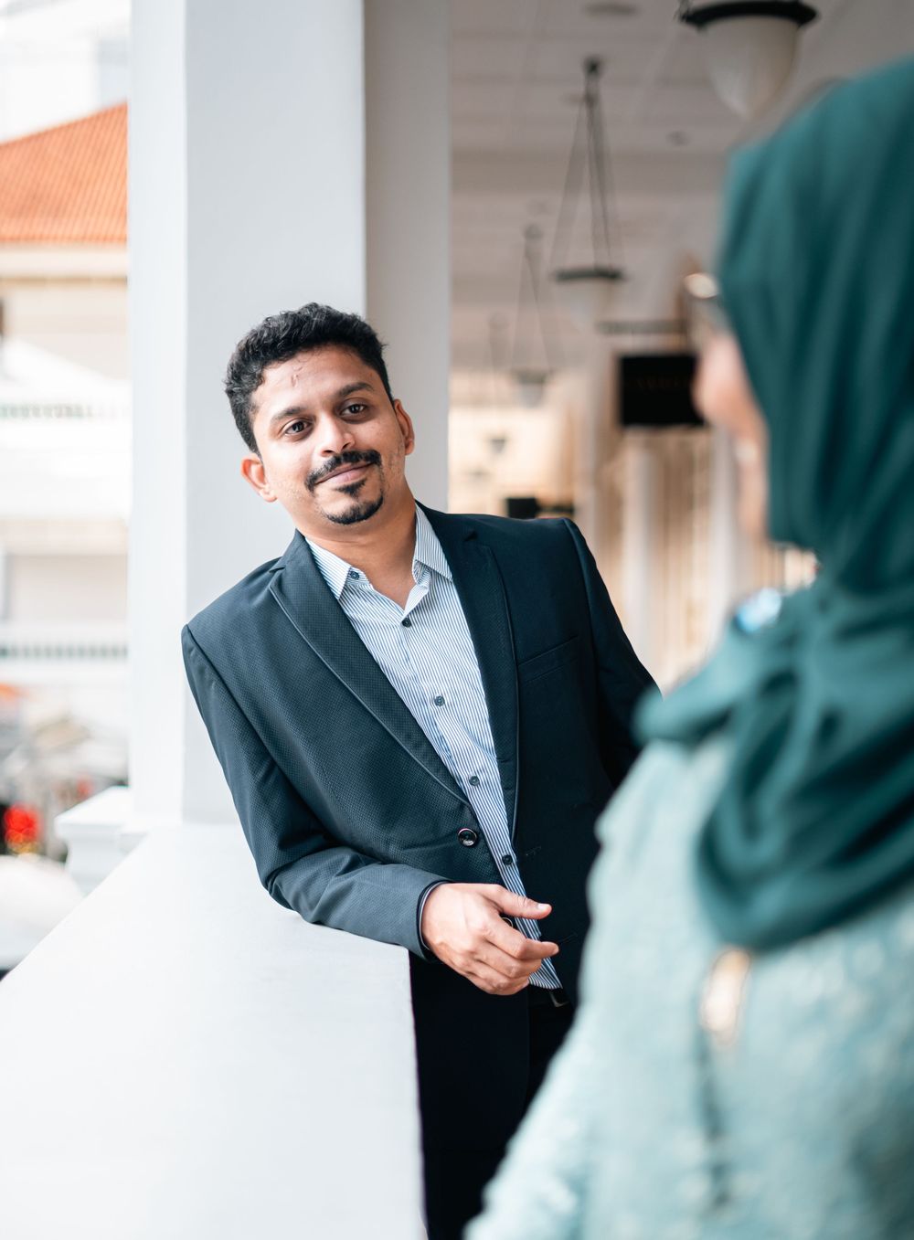 A man in a dark blazer smiles while conversing with a woman wearing a green hijab in a bright indoor setting.