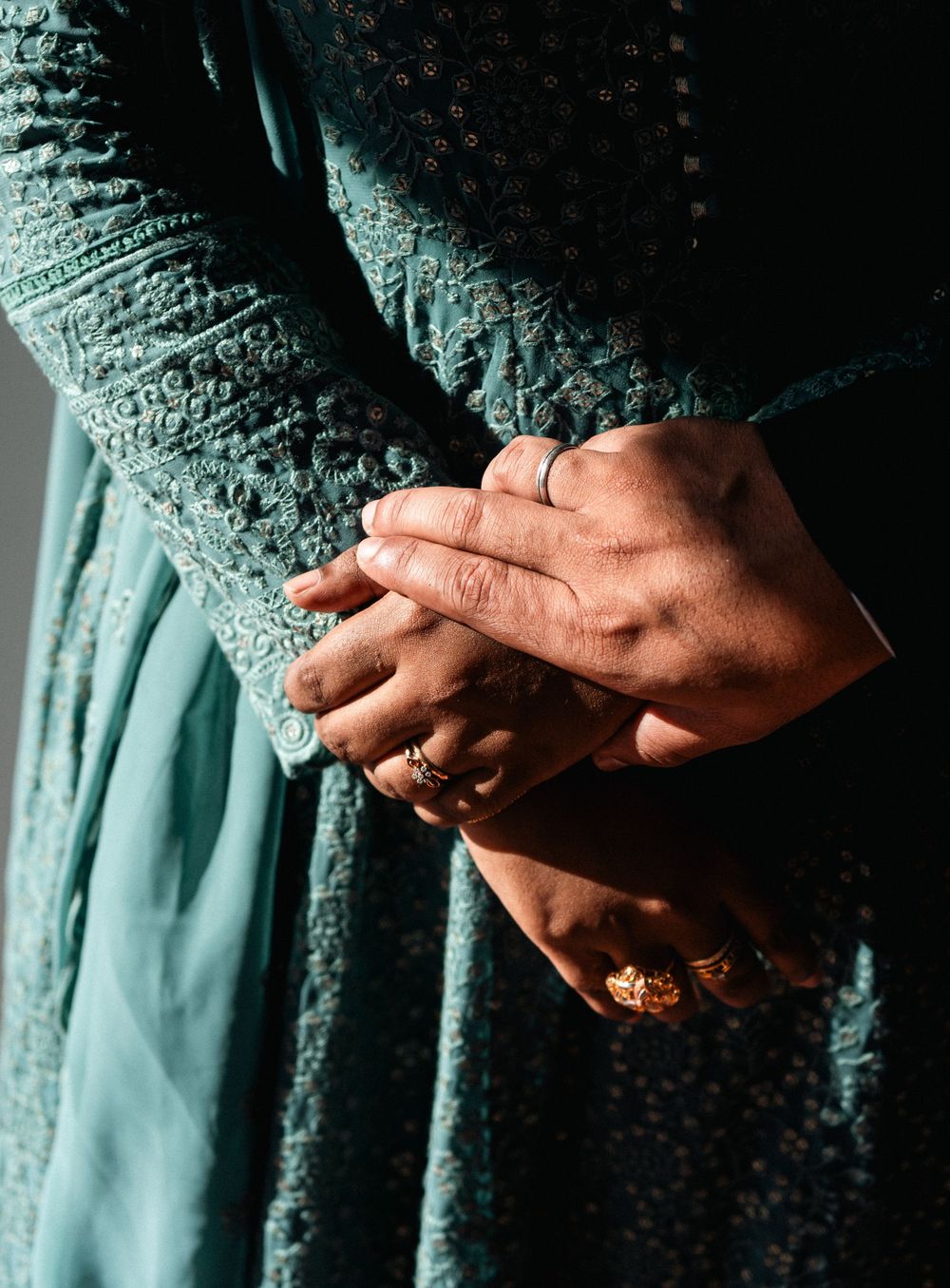 Close-up of clasped hands adorned with gold and silver rings, wearing a teal embroidered lace dress.
