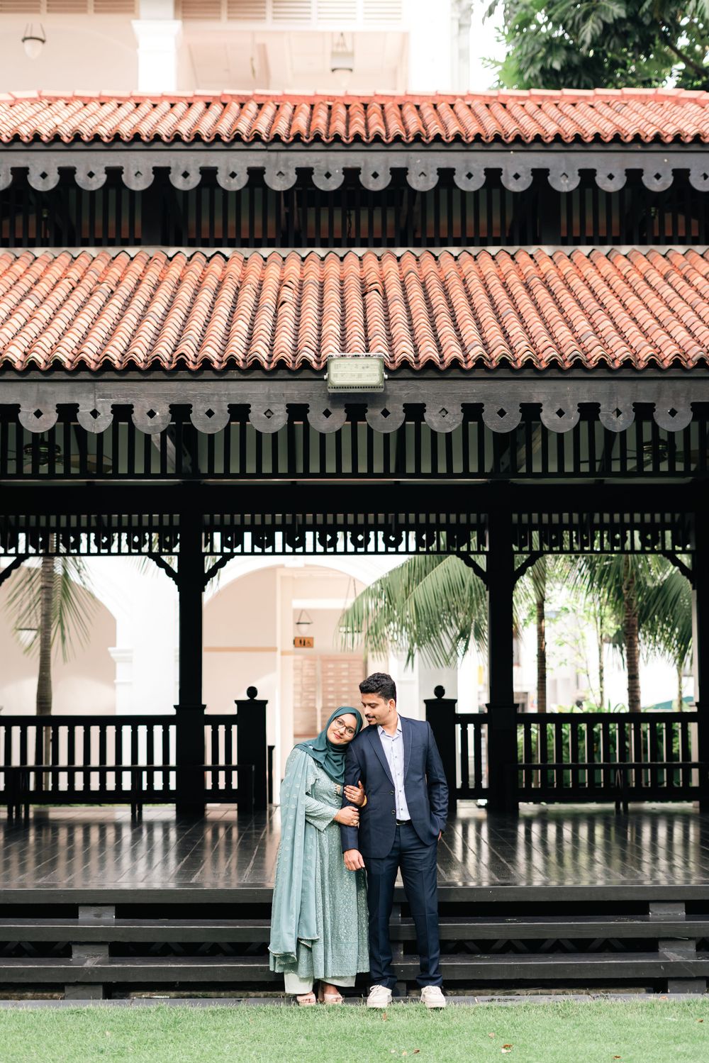 Muslim couple in elegant attire poses under a traditional wooden pavilion with red tiled roof.