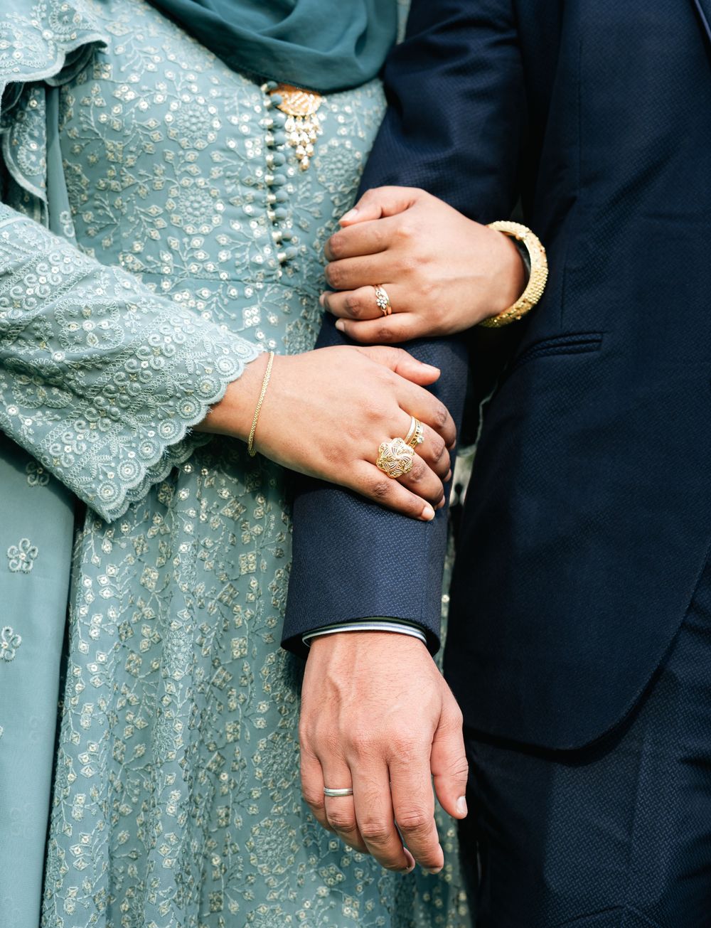 Close-up of couple's hands adorned with gold rings and bracelets, wearing blue embroidered dress and suit.