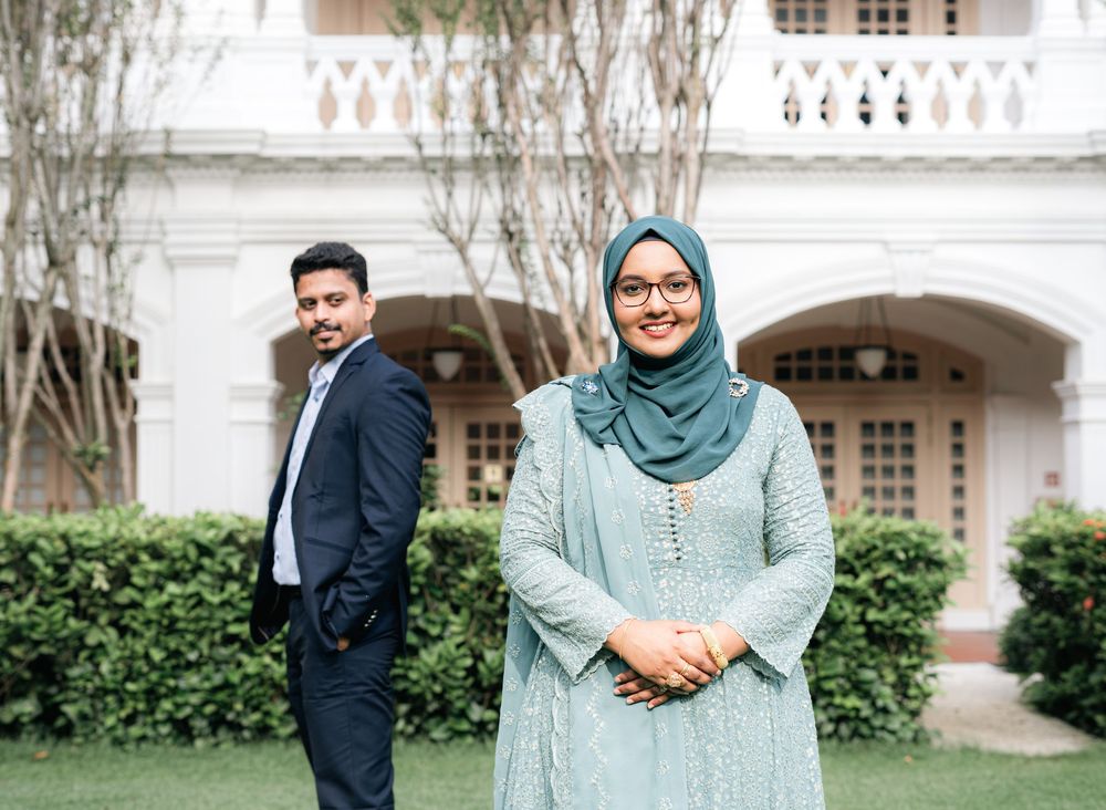 A man in a navy suit and a woman in a teal hijab and embroidered dress pose outdoors near a white building.