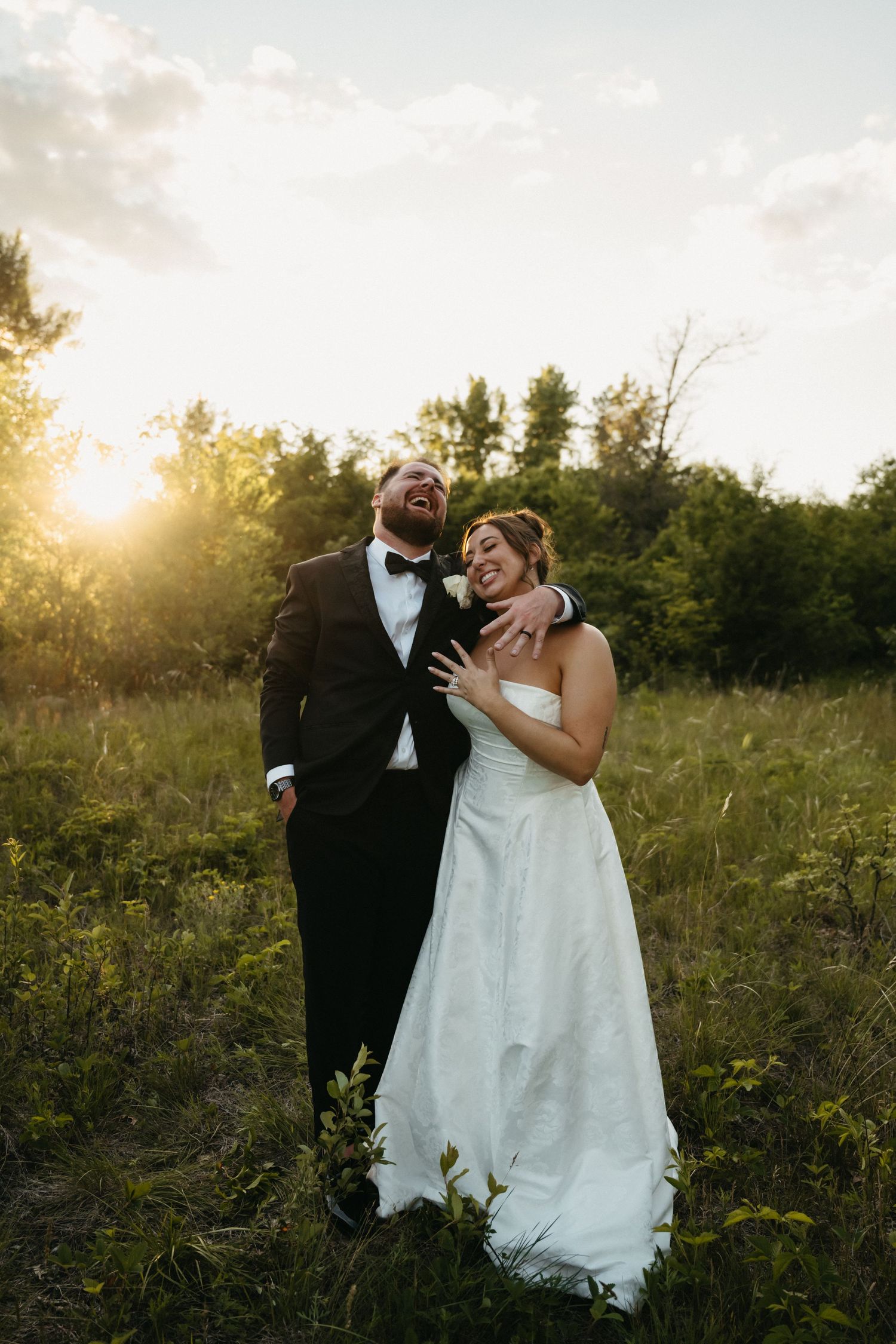 Joyful bride and groom laughing together in a sunlit green field during golden hour wedding portrait.