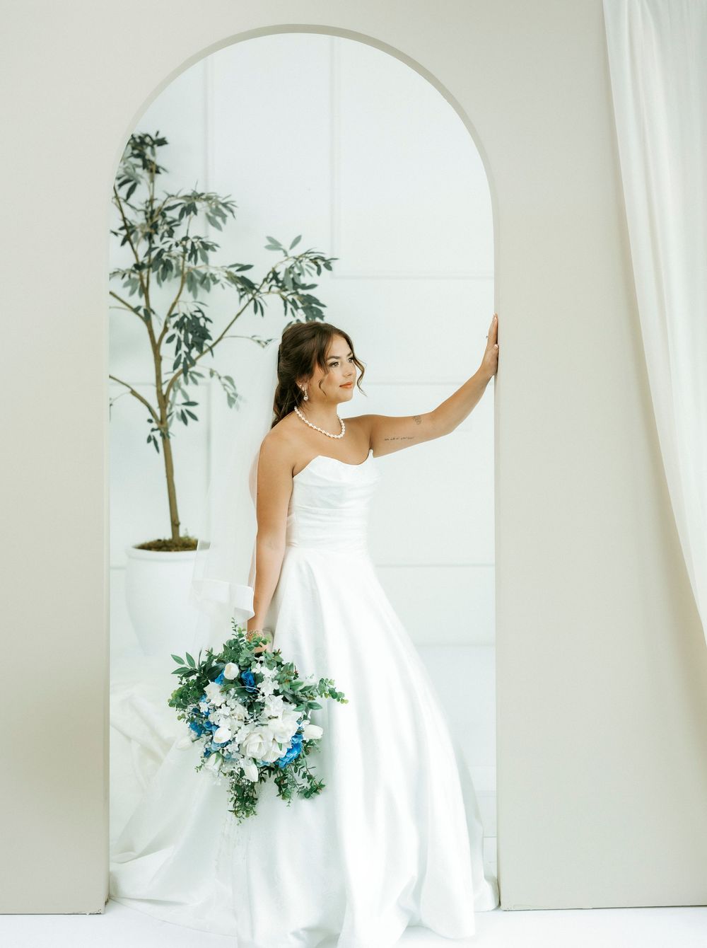 Bride in strapless white gown holds blue and white bouquet beside arched mirror with potted tree.