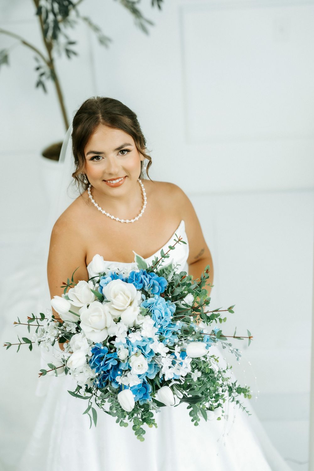 Bride in white strapless gown holding blue and white floral bouquet with pearl necklace smiling indoors.