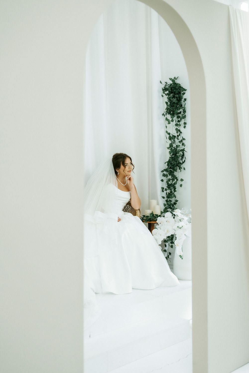 Bride in white wedding gown and veil sits gracefully in bright white room adorned with lush green ivy.