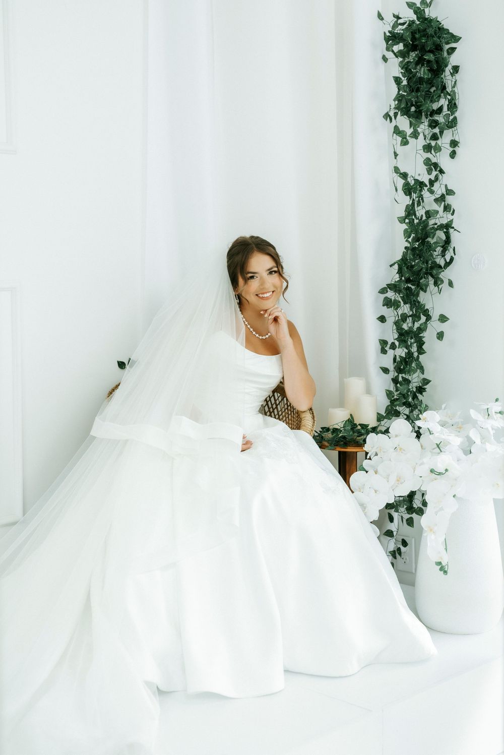 Smiling bride in a white ball gown and veil sits gracefully in a bright white room with green ivy and white flowers.