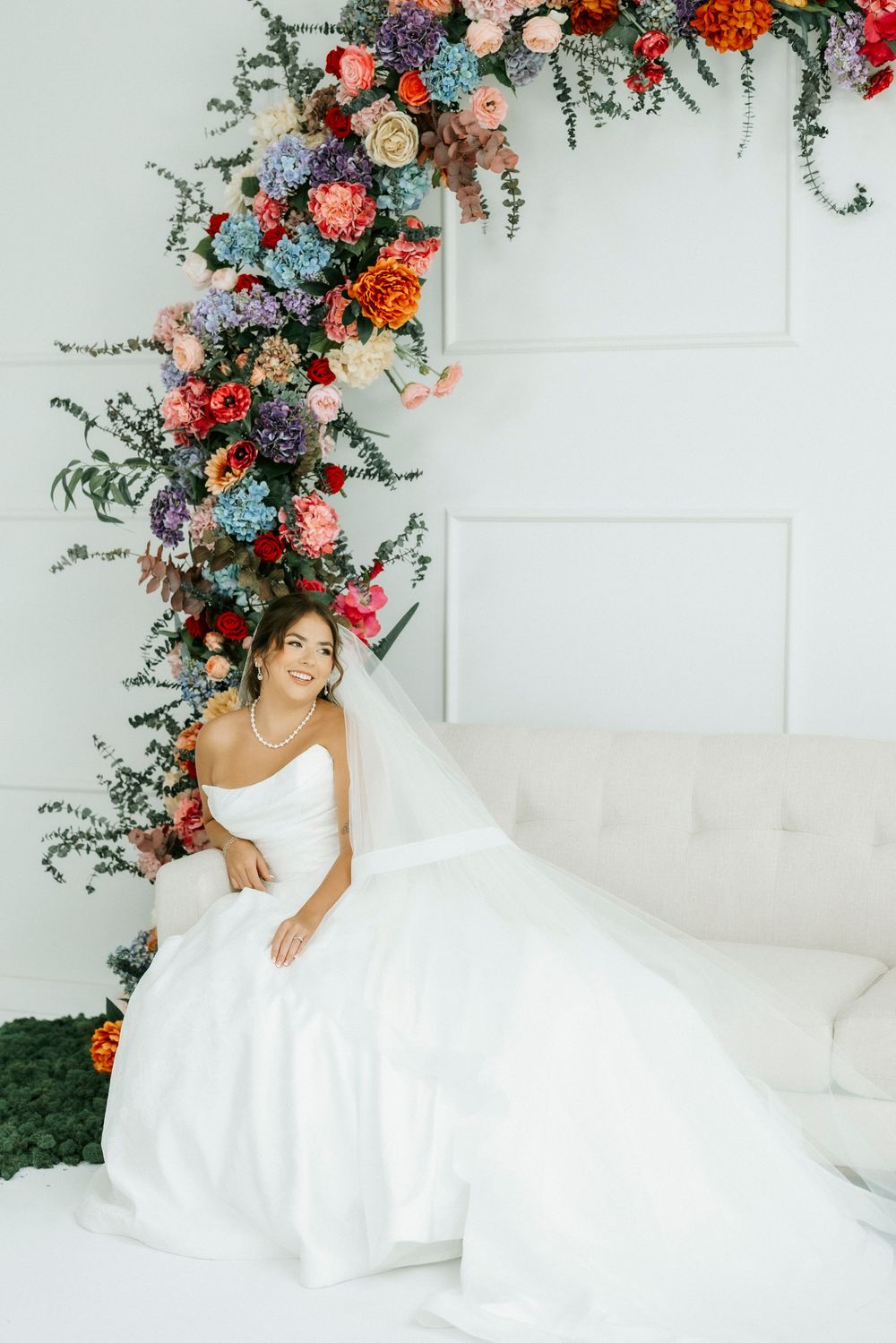 Bride in strapless white gown sits on sofa beside colorful floral arch with roses and greenery in bright white studio.