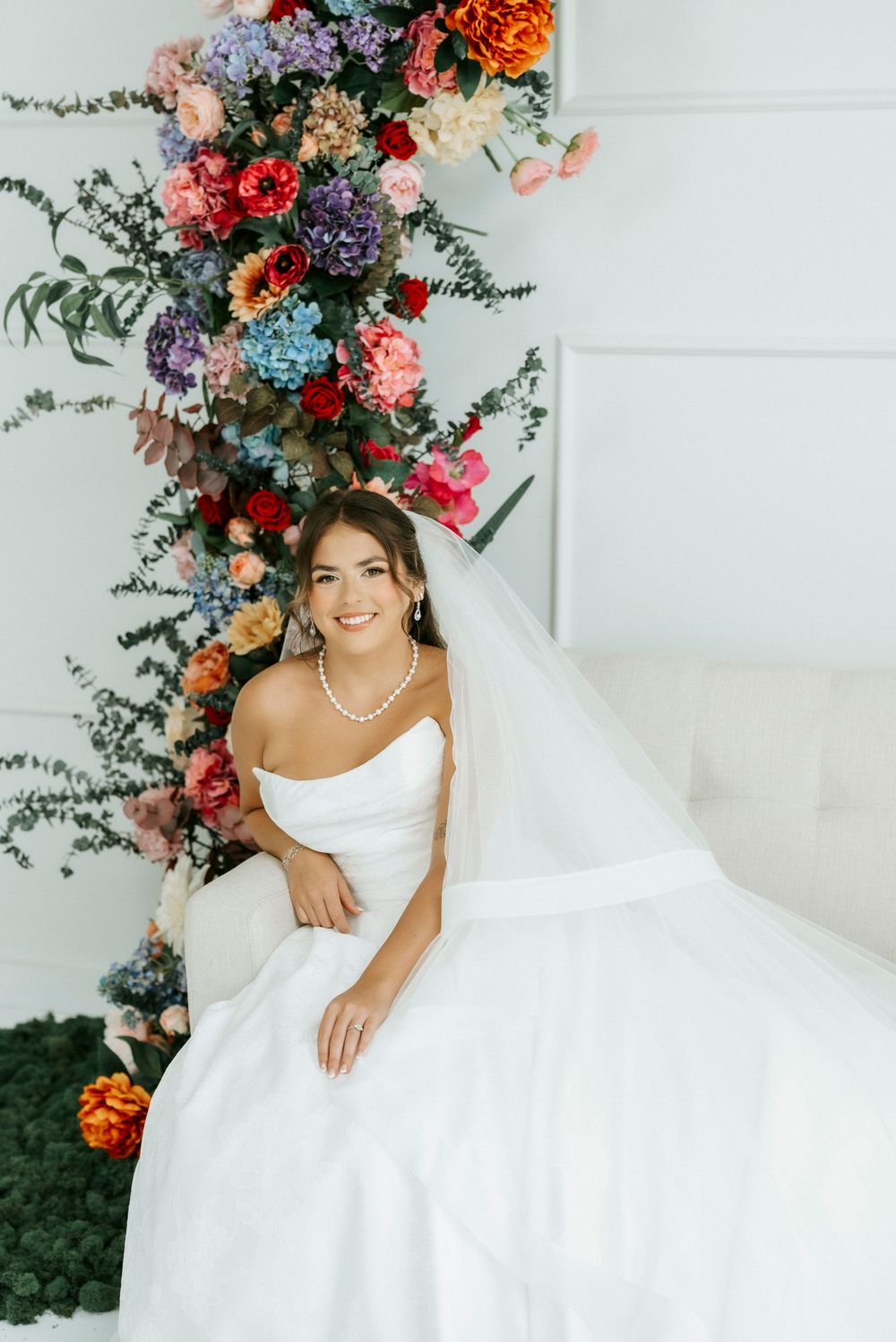 Bride in white strapless gown with veil sitting before colorful floral arrangement wearing pearl necklace.