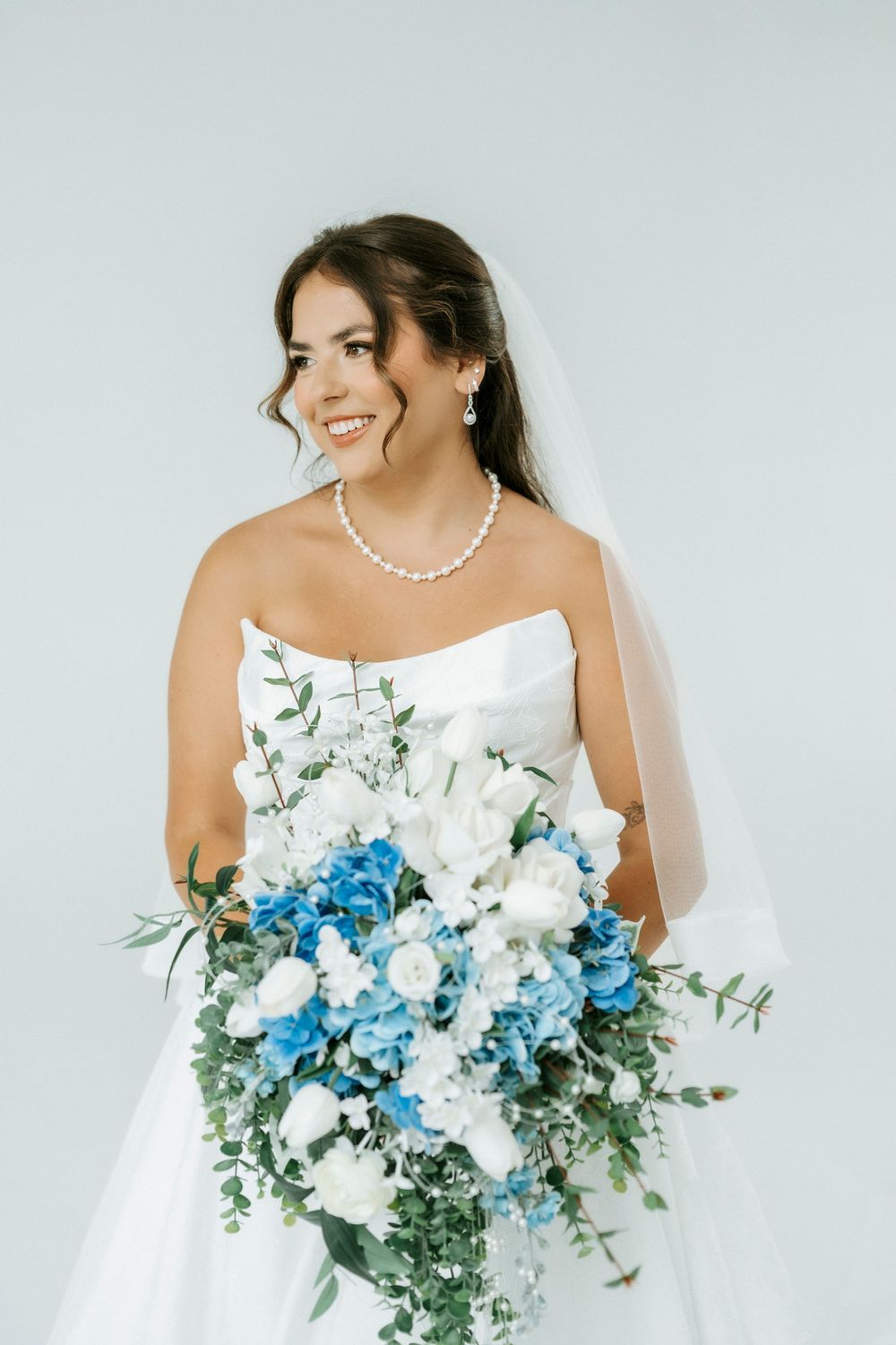Smiling bride in strapless white gown holding cascading blue and white floral bouquet with pearl necklace and veil.