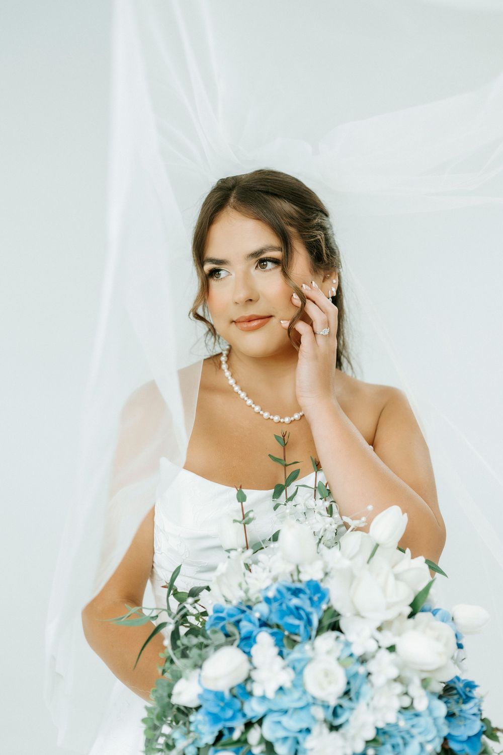 Bride in white strapless gown holding blue and white floral bouquet, wearing pearl necklace, posing under flowing veil.