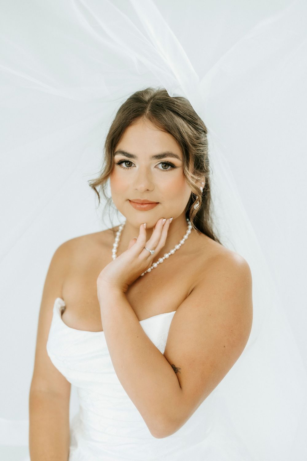 Elegant bride in white strapless gown with pearl necklace and earrings, posing gracefully under a flowing veil.