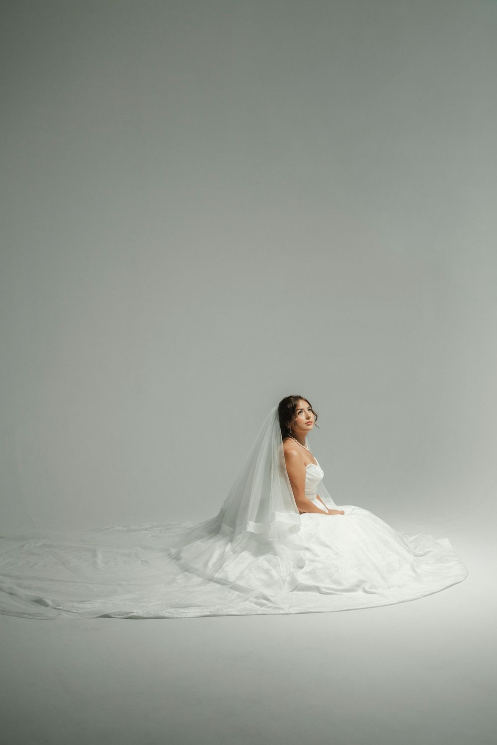 A bride in a white strapless gown and long veil sits gracefully on the floor against a soft gray background.