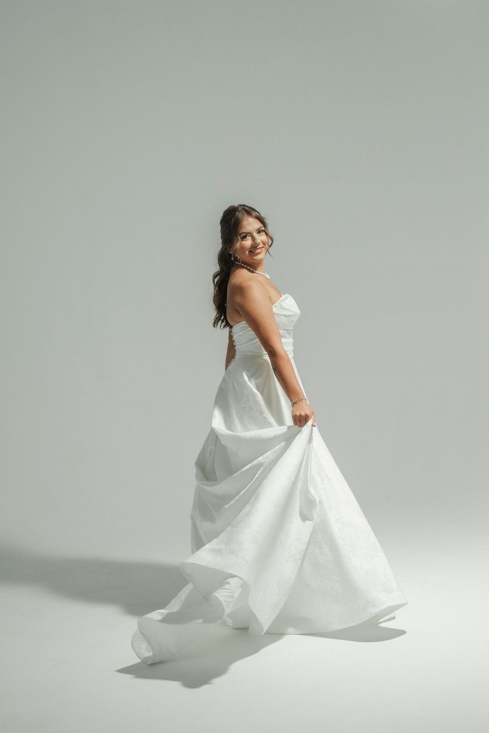 A smiling bride twirls in a flowing strapless white wedding gown against a soft gray studio background.