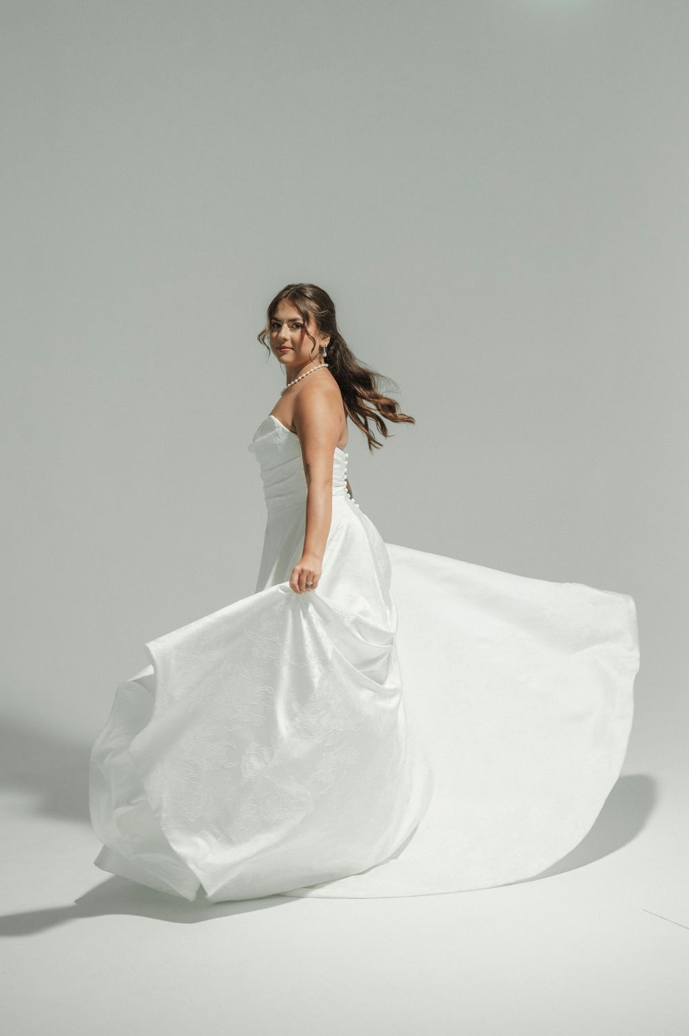 Bride twirls in flowing white strapless wedding dress, smiling over shoulder on light gray background.