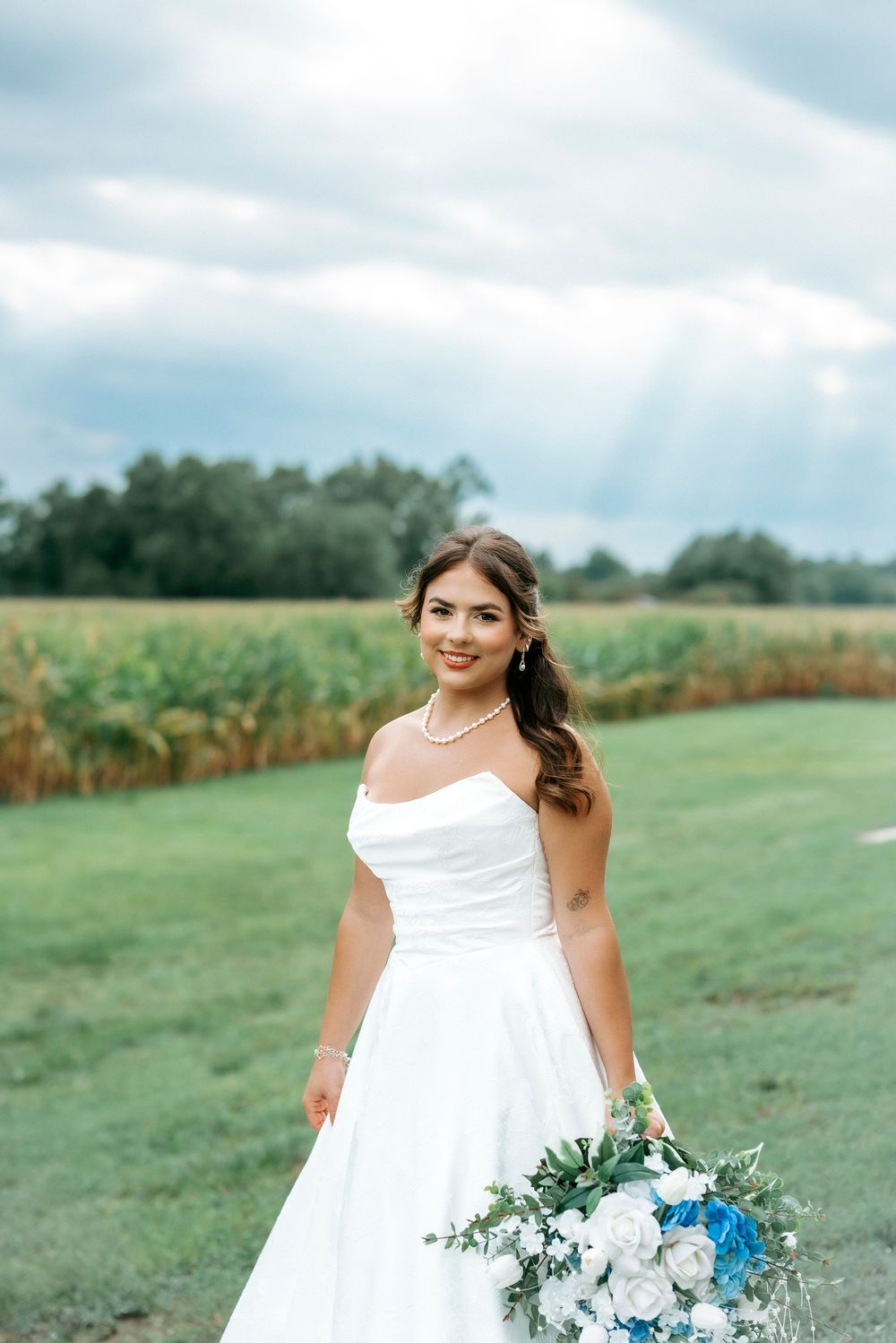 Smiling bride in strapless white ball gown holds blue and white bouquet in front of cornfield.