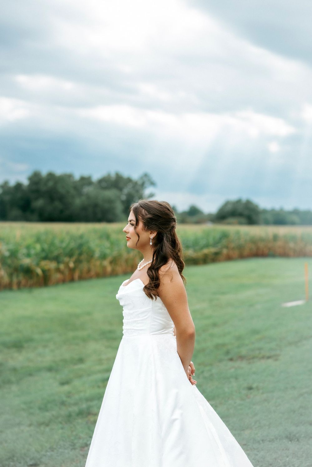 Bride in strapless white gown twirls gracefully in green field with cornfield and cloudy sky backdrop.