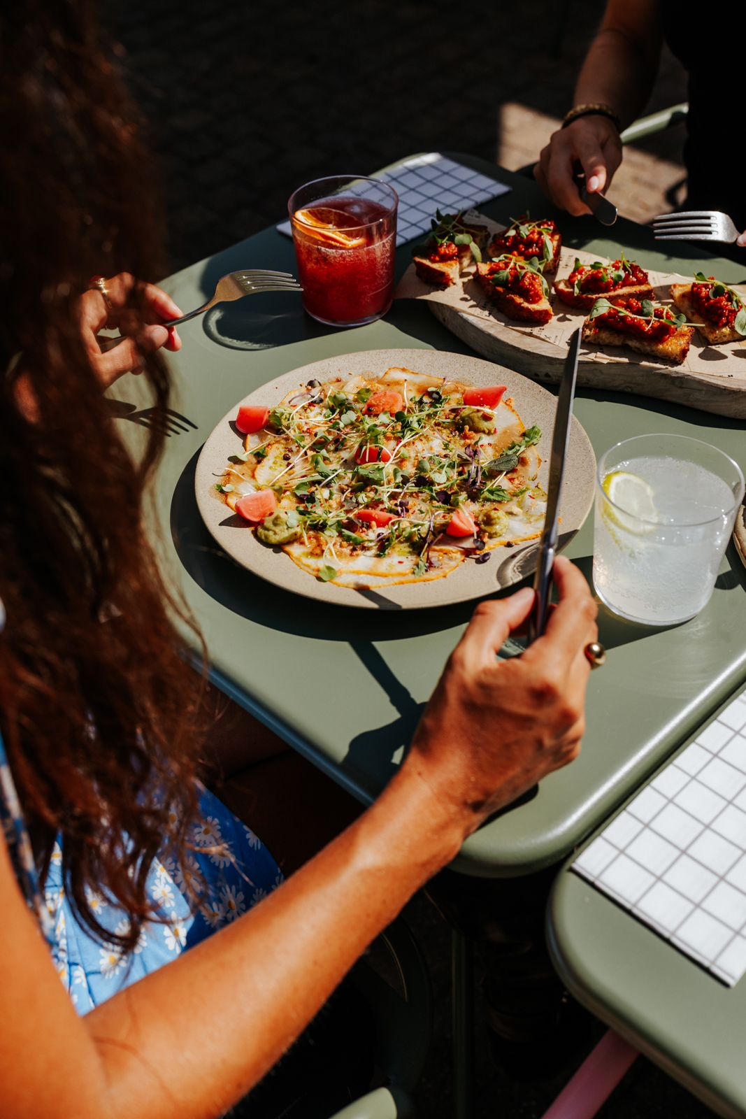 Woman cutting fresh pizza with knife and fork at outdoor cafe table with drinks and bruschetta in sunlight.