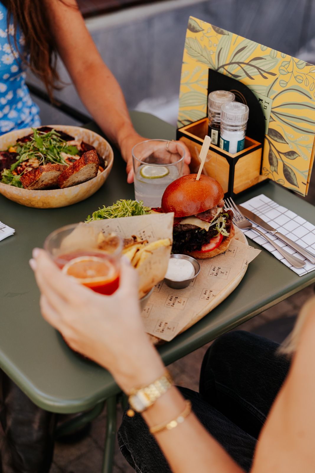 Two people dining outdoors with a burger, salad bowl, and drinks on a green table at a casual restaurant.