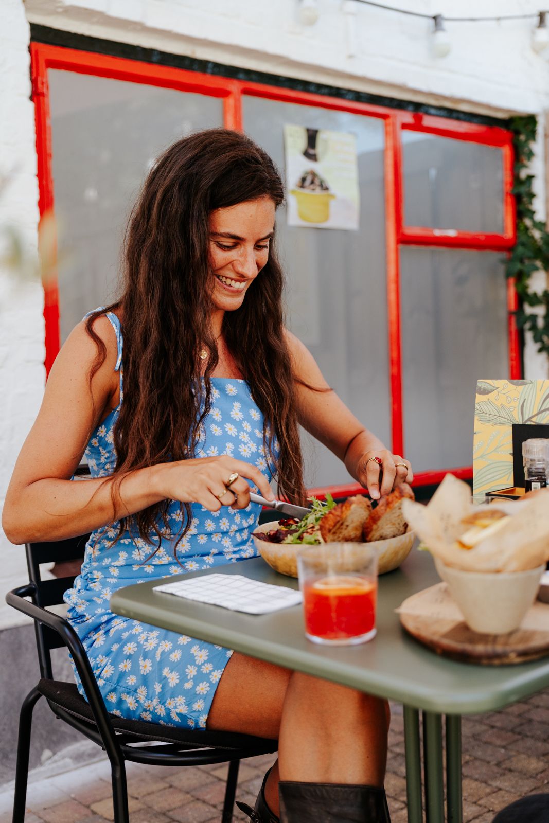 Smiling woman in blue floral dress dining outdoors at cafe table with salad and red drink.