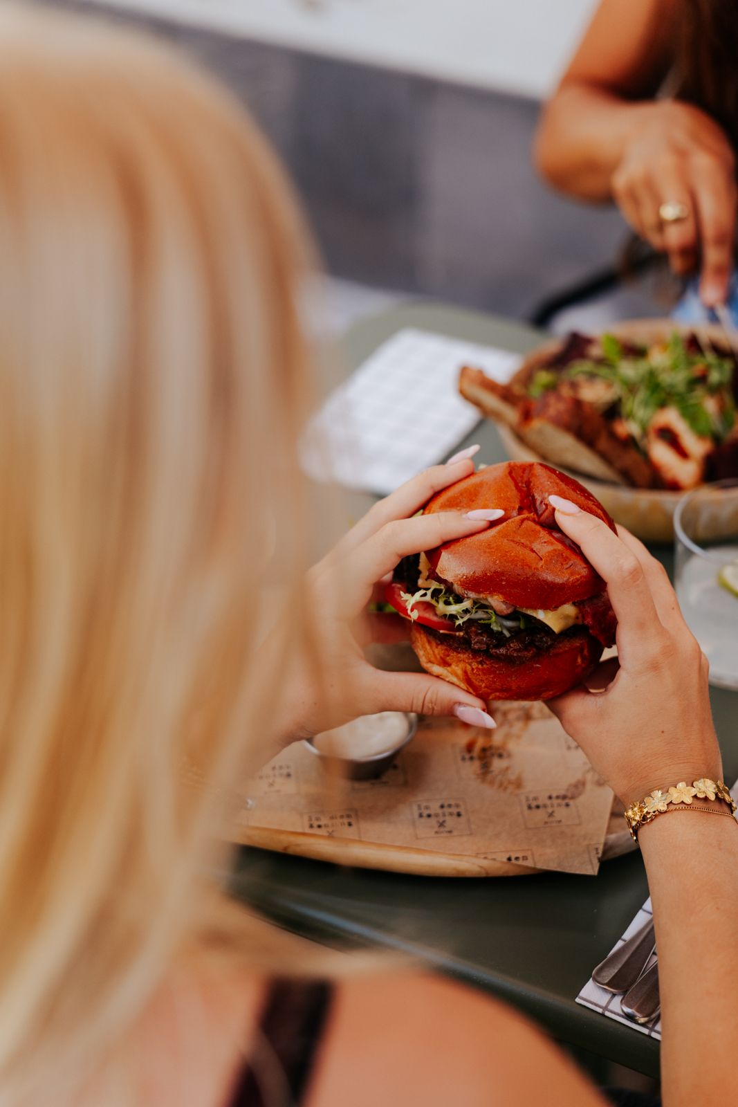 A blonde woman holds a juicy burger with a red brioche bun over parchment paper at a casual dining table.