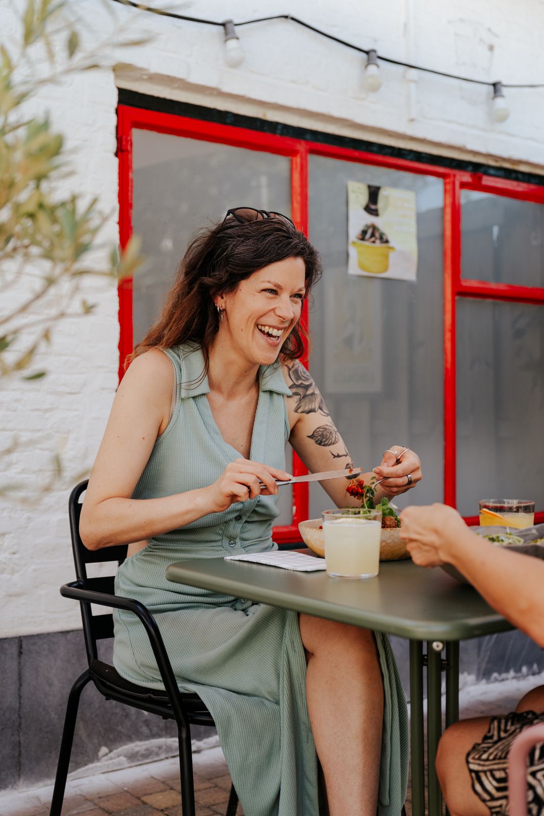 A laughing woman in a sage green dress with tattoos enjoys a meal at an outdoor cafe table with string lights.