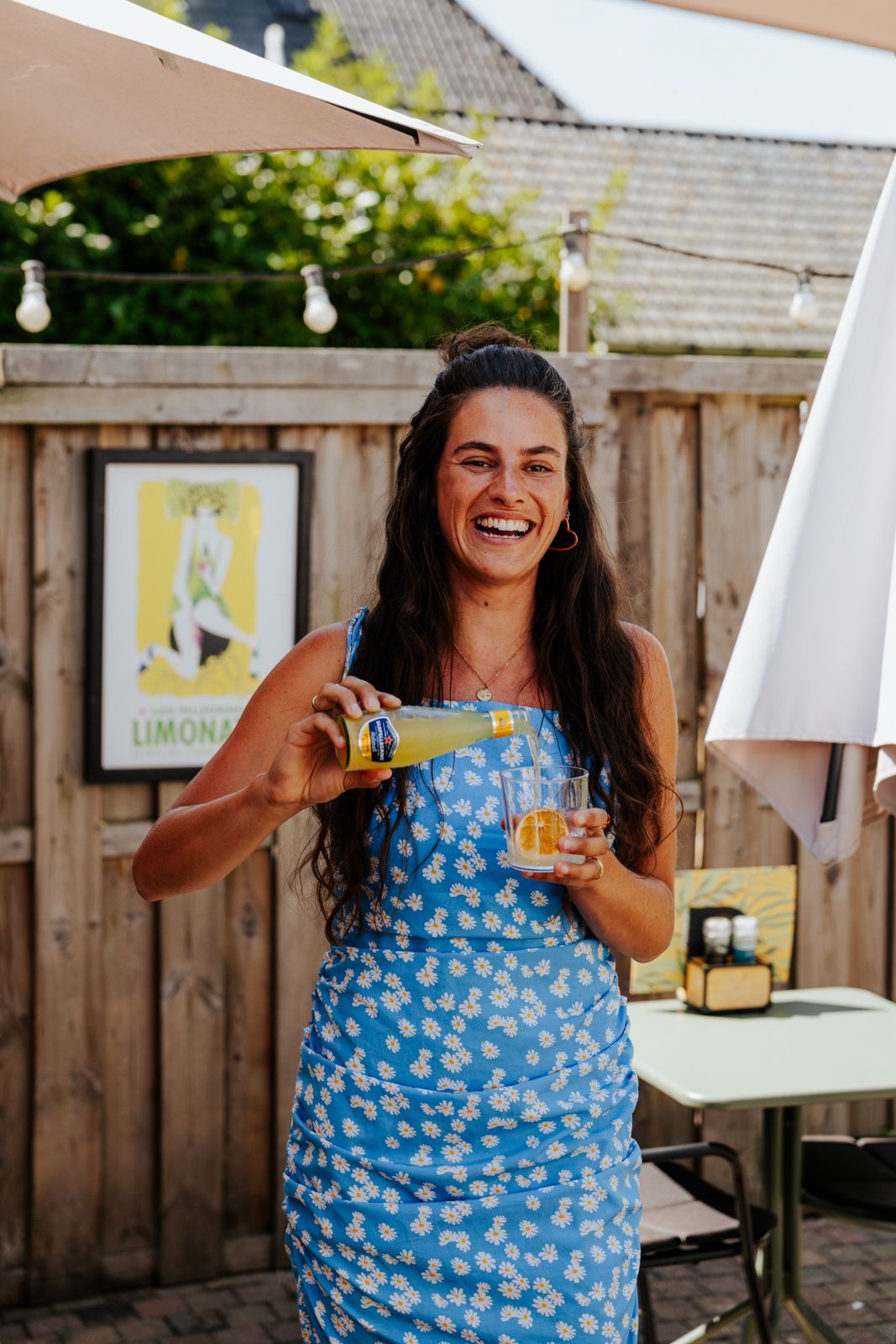 Smiling woman in blue floral dress holding lemonade bottle and glass in sunny backyard garden setting.