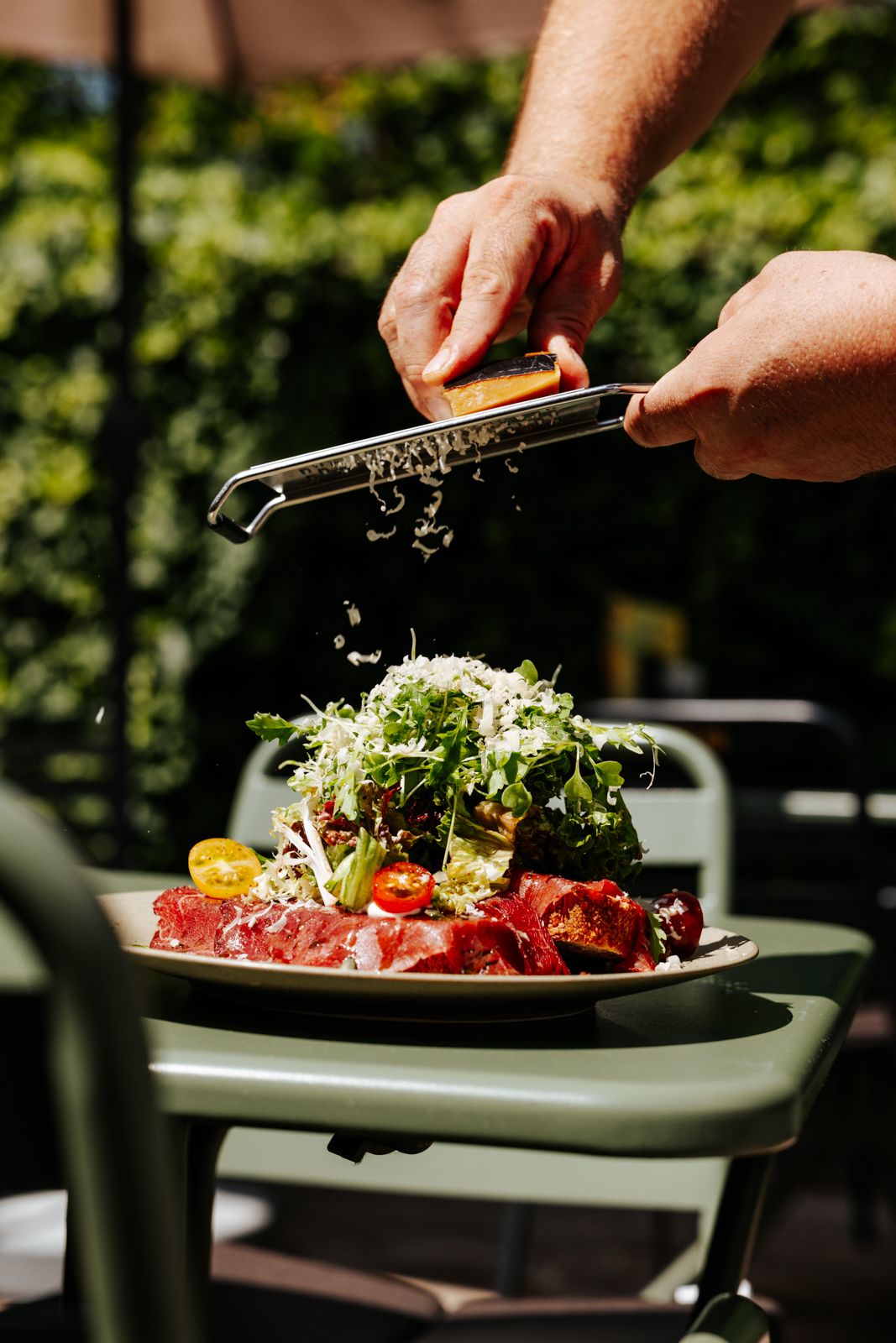 Chef grating fresh cheese over a gourmet steak salad with arugula and cherry tomatoes on an outdoor patio.