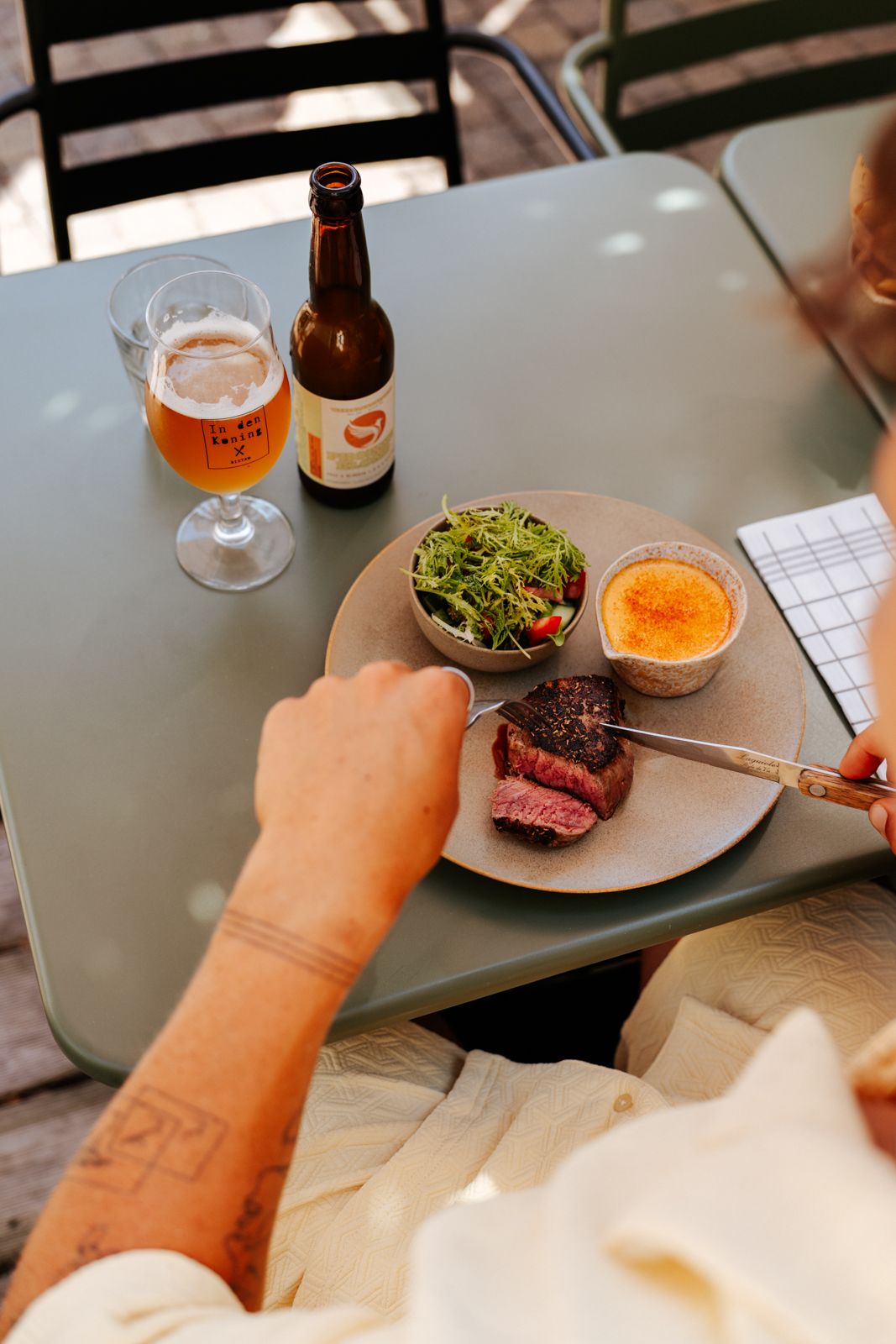 Person cutting a medium-rare steak on a plate with salad and gratin, beside a craft beer bottle and glass.