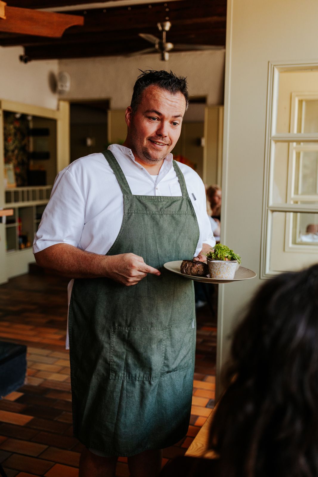 Chef in green apron presenting a gourmet plate with meat and greens to a restaurant guest.