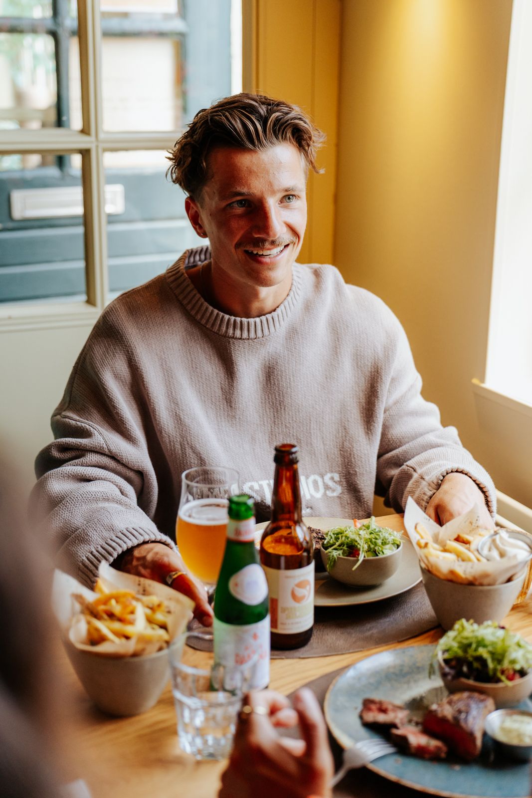 Smiling man in beige sweater enjoying lunch with beer, fries, and salad at a bright restaurant table.