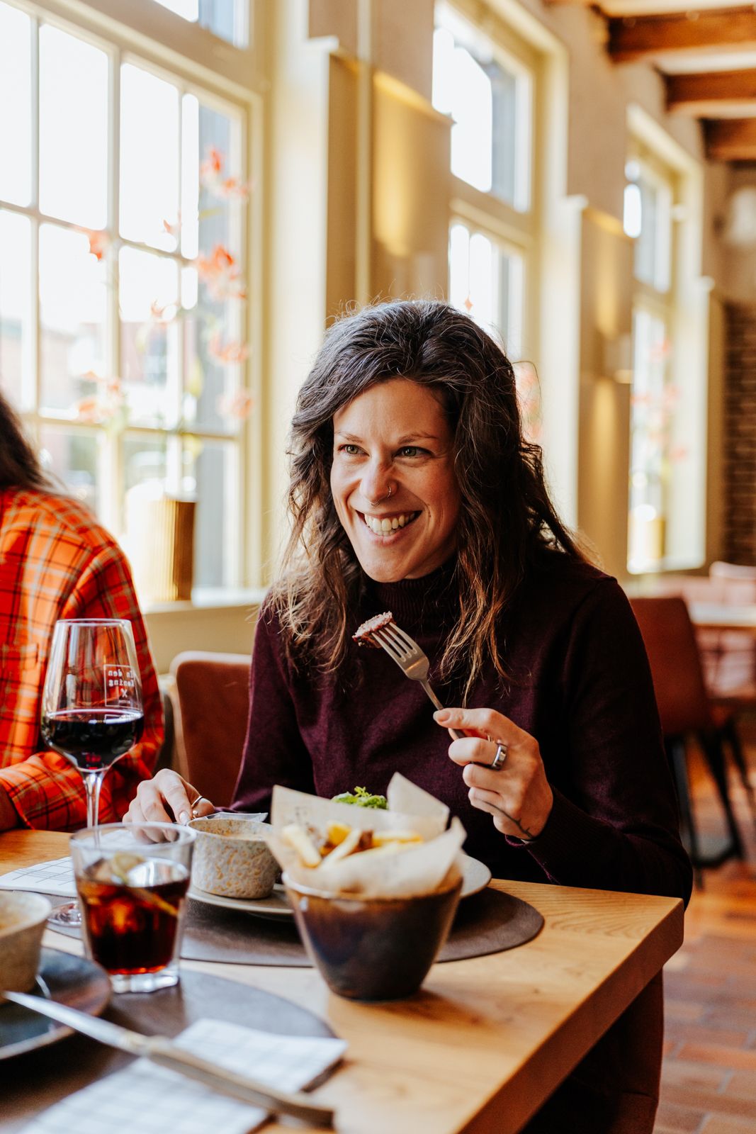 A smiling woman enjoys a meal with wine at a cozy restaurant, holding a fork while dining with a friend.