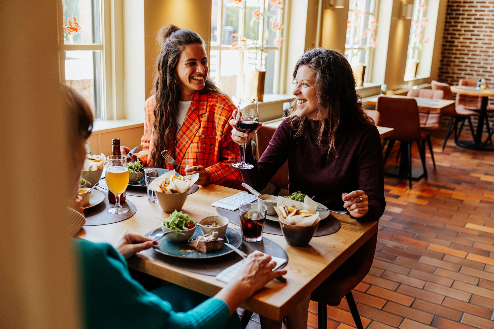 Three women laughing and toasting with wine and juice at a restaurant table with plates of food.
