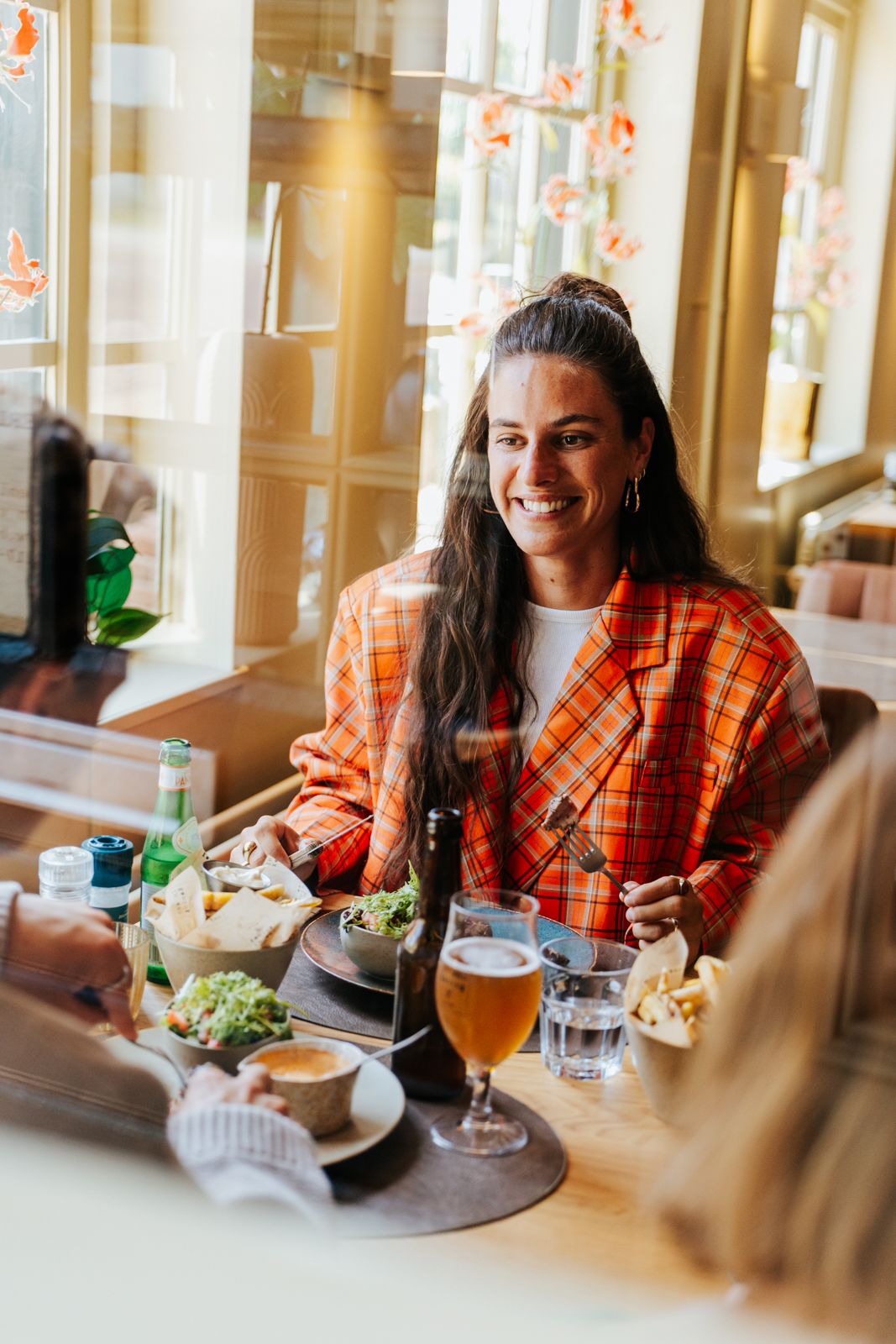 Smiling woman in orange plaid blazer enjoying lunch with beer at a cozy restaurant table.