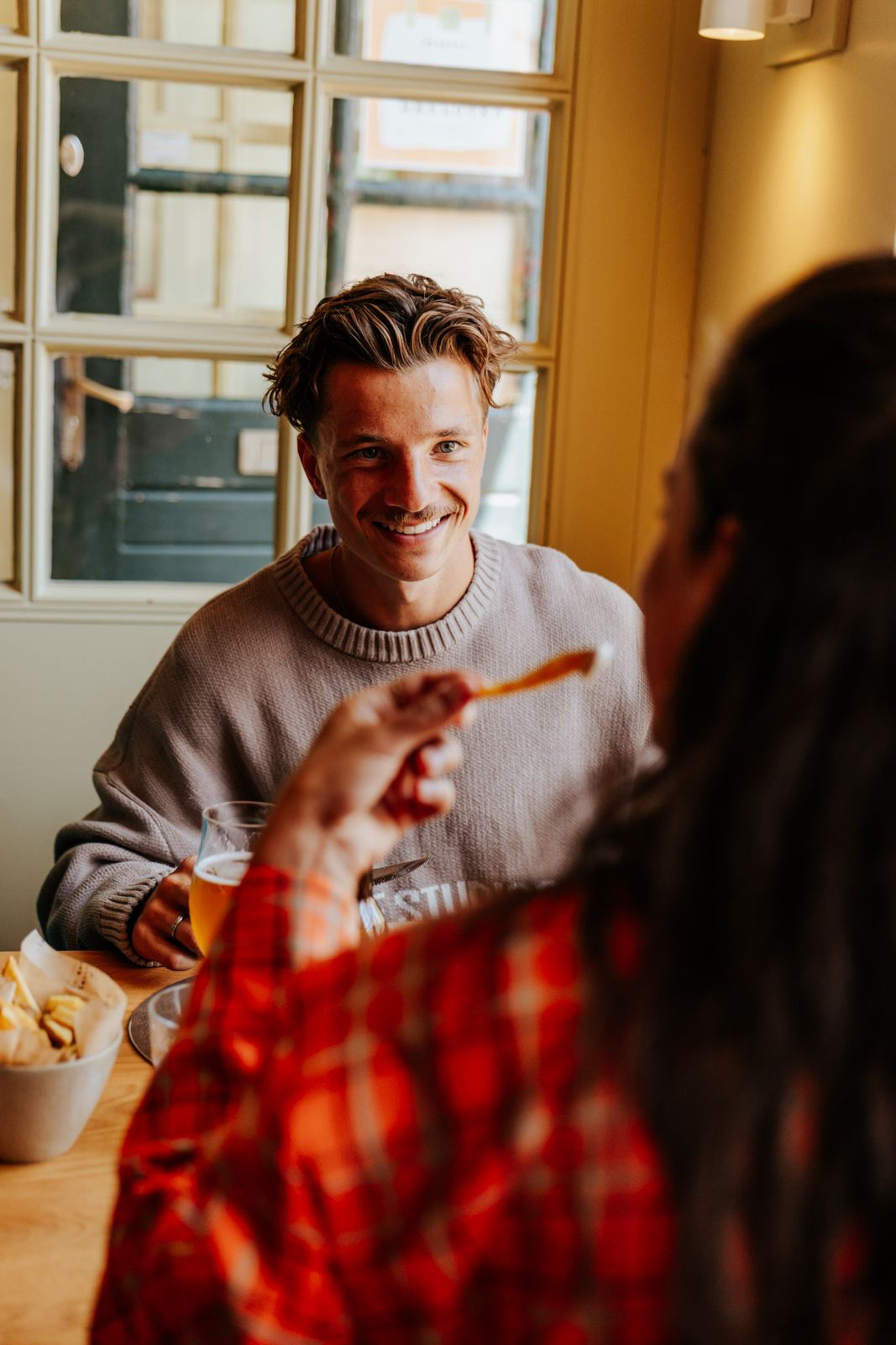 Smiling young man in gray sweater enjoying a meal with a friend at a cozy restaurant table near a window.