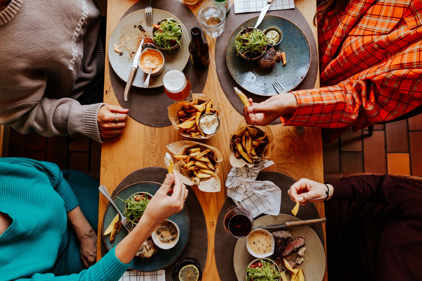 Overhead view of friends sharing a meal with fries, steak, and salads on a wooden table.