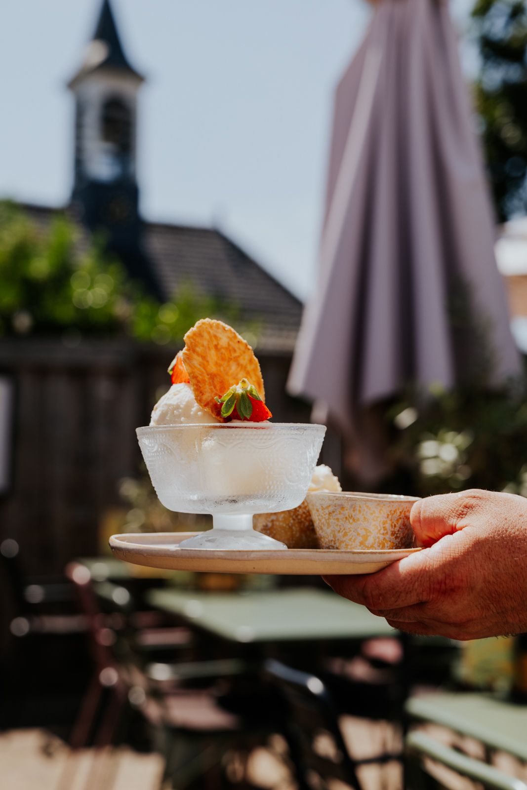 Hand holding a plate with an elegant ice cream sundae topped with a wafer cookie, served outdoors at a charming cafe.