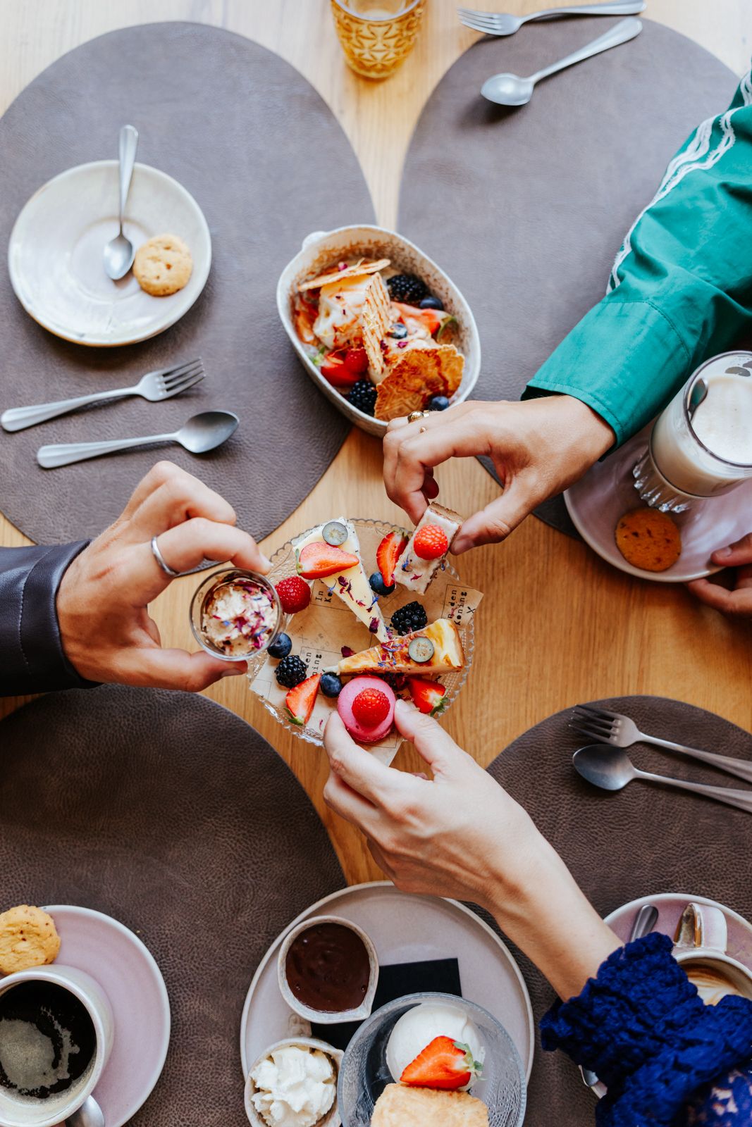 Overhead view of people sharing desserts with fresh berries, coffee, and drinks at a round wooden restaurant table.