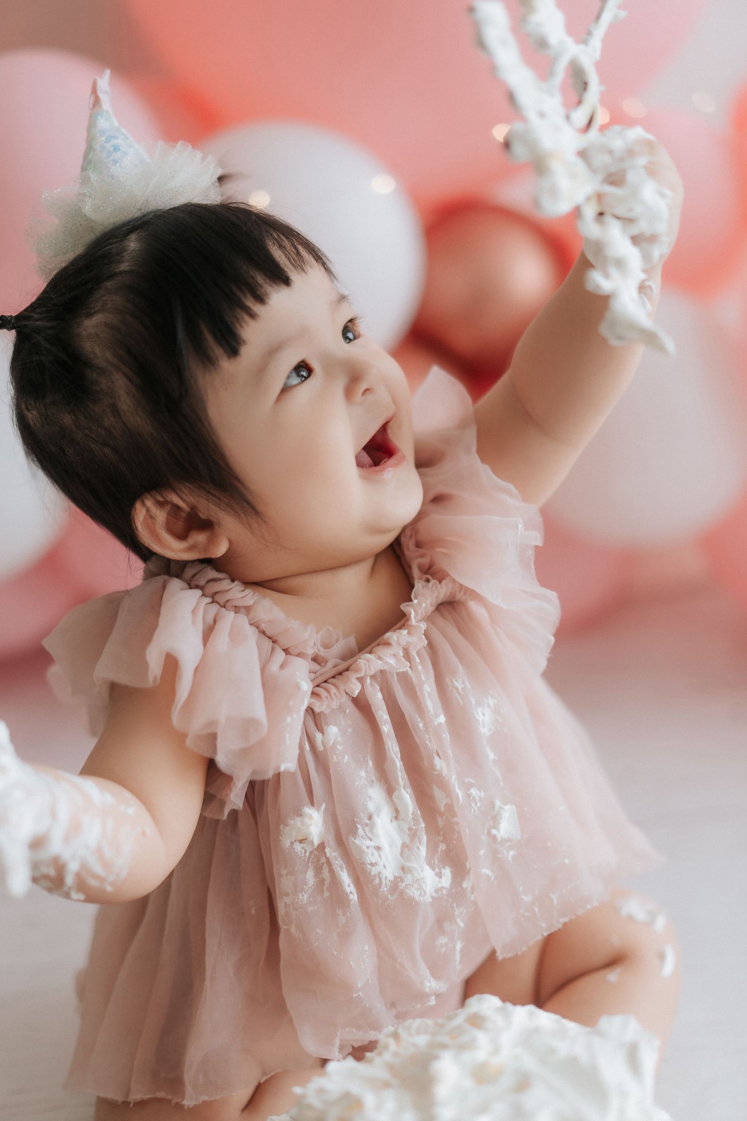 A joyful baby girl in a pink tutu dress celebrates her first birthday smashing cake, surrounded by pink balloons.