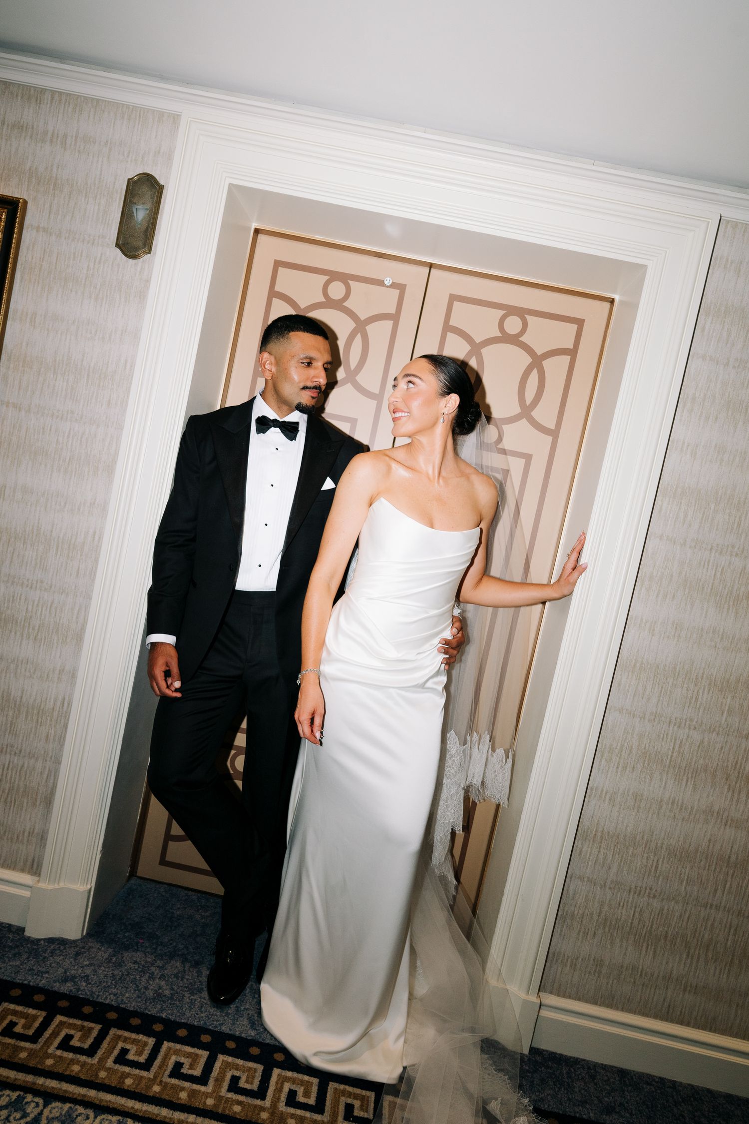 Elegant bride in strapless white gown and groom in black tuxedo sharing a romantic moment by ornate gold doors.