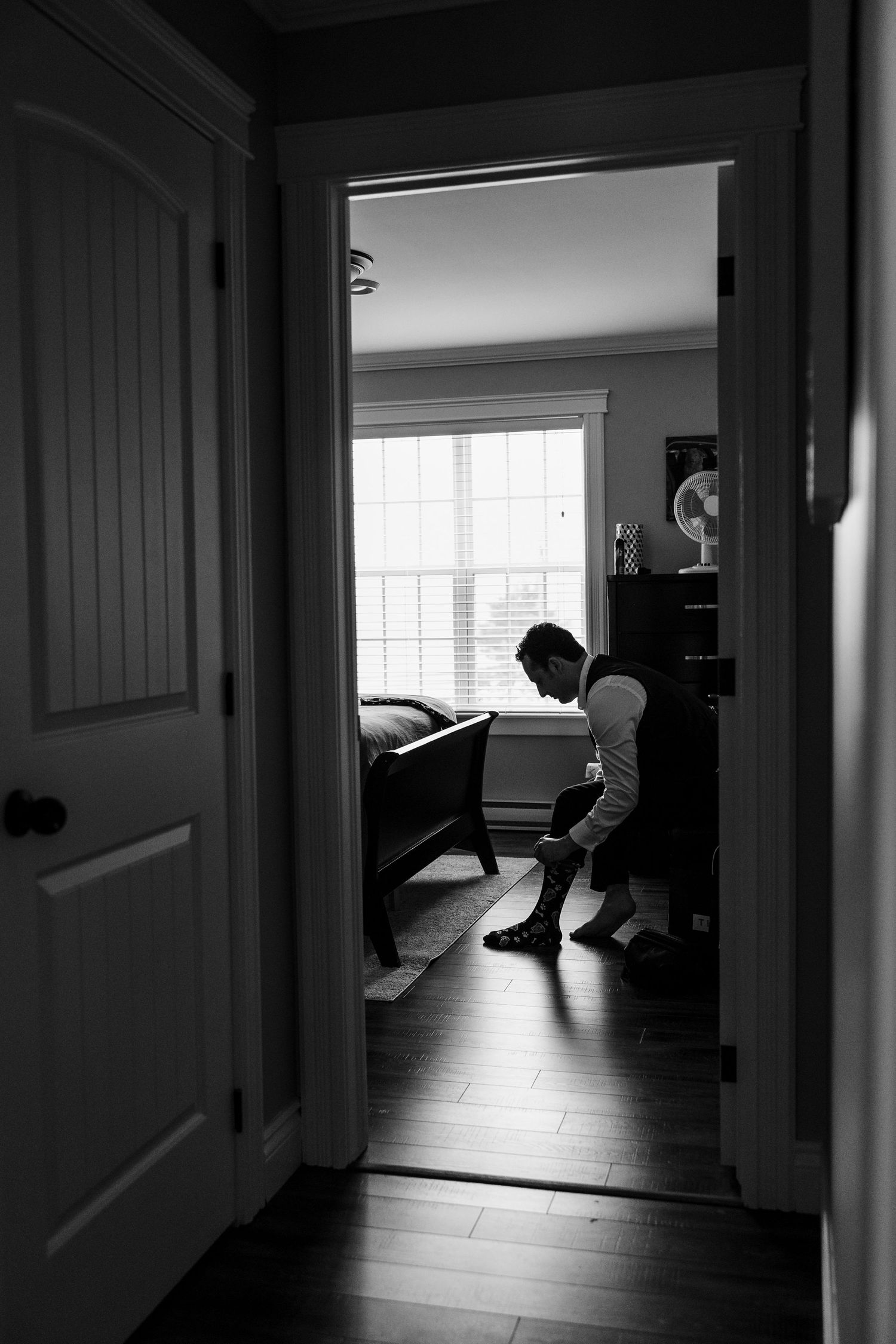 Black and white photo of a person getting dressed in a sunlit bedroom, viewed through a doorway.