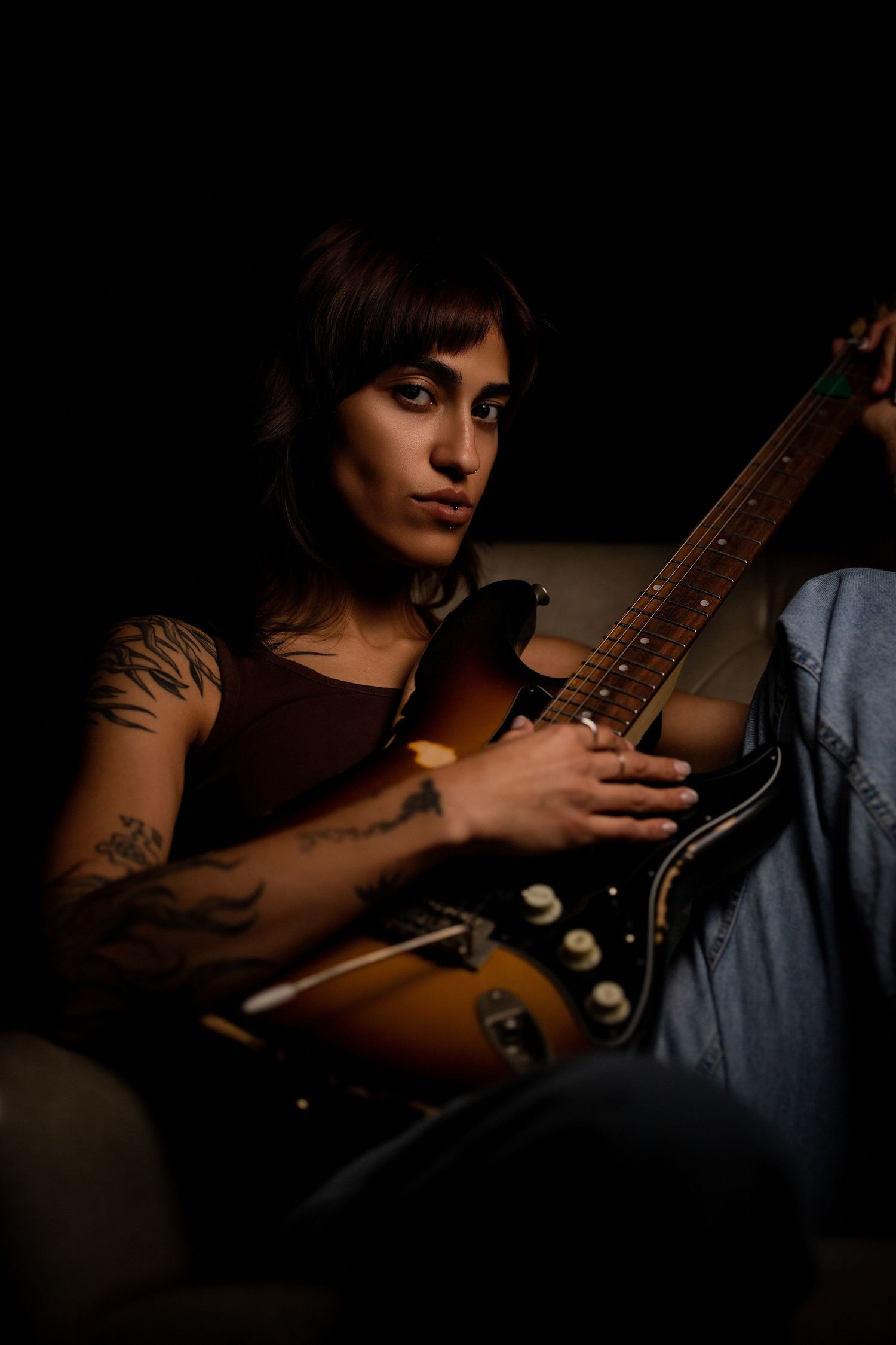 Tattooed woman with dark hair holding a sunburst electric guitar in dramatic low light.