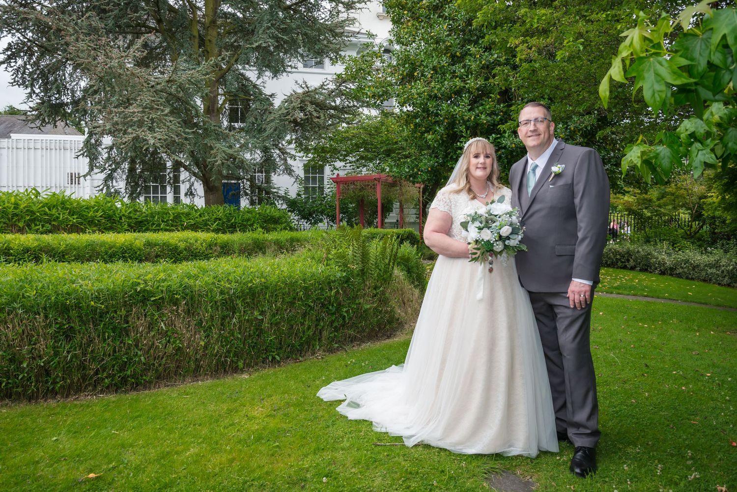 Black and white wedding photo of bride and groom standing in elegant room with large windows and family seated nearby.