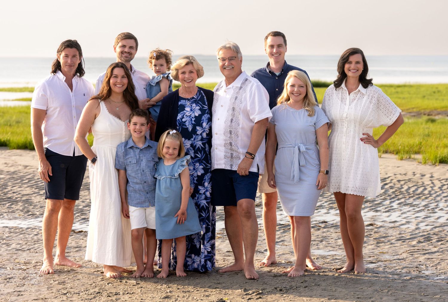 Large family group portrait smiling together on a sandy beach at golden hour with marsh grass and ocean in background.