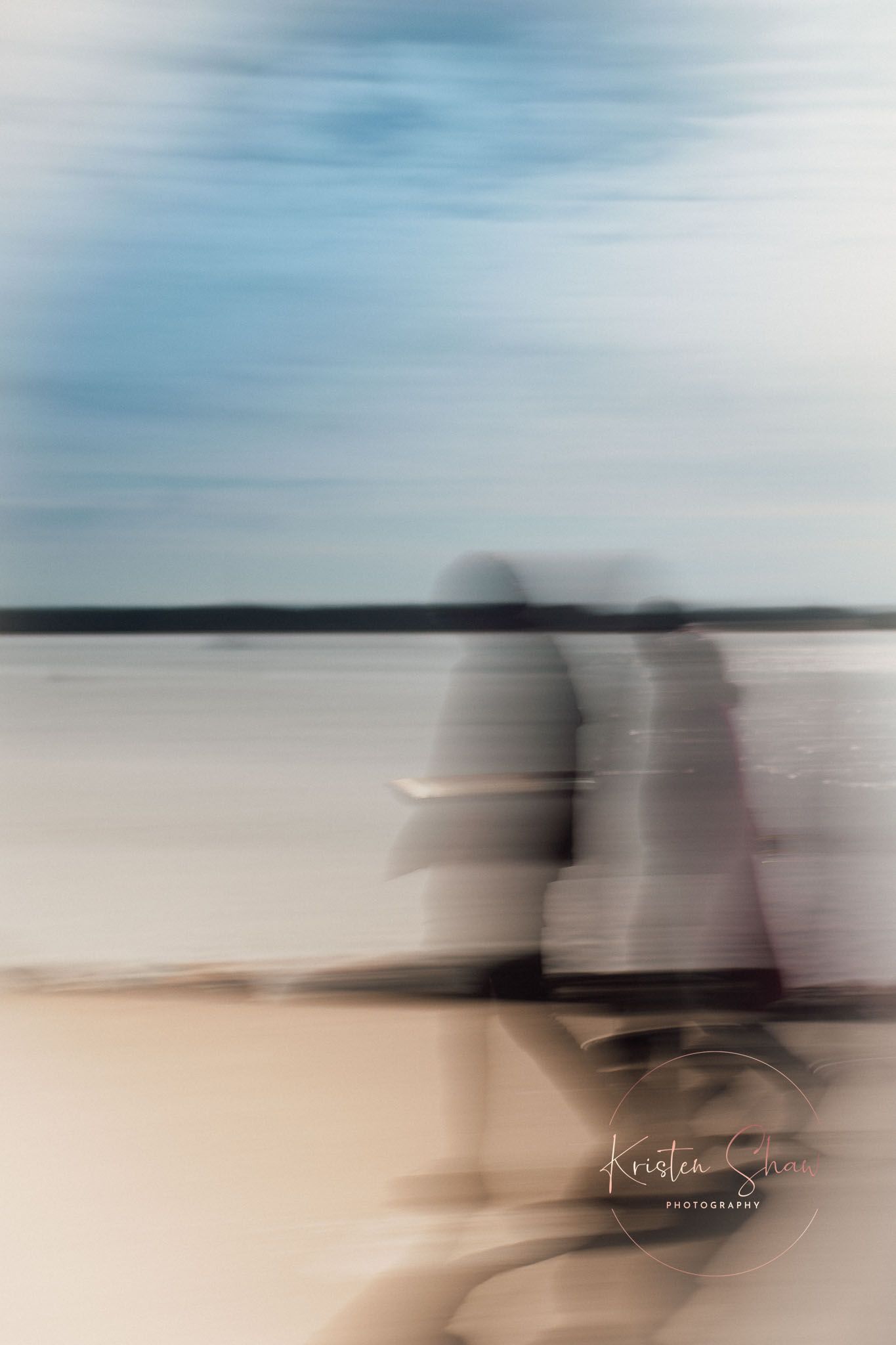 Blurred motion photo of two people walking along a serene lakeside beach with a soft blue sky.