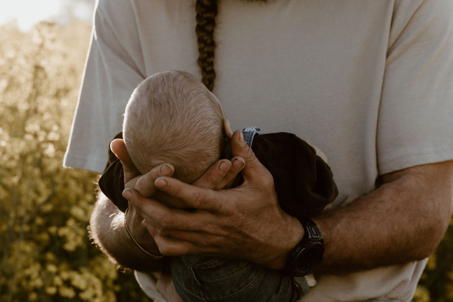 A bearded man in a white t-shirt tenderly cradling a newborn baby outdoors in natural light.