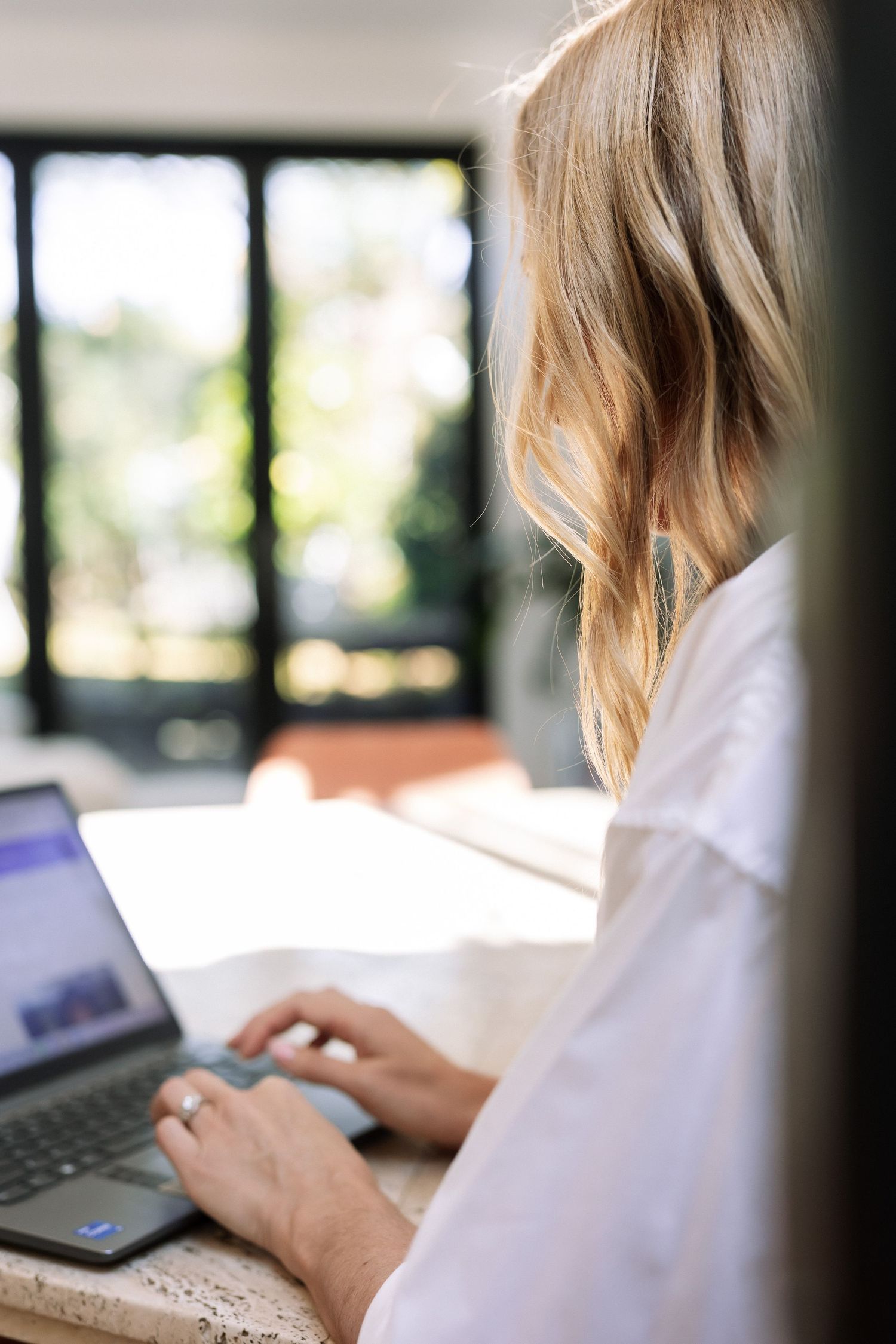 Blonde woman in white shirt working on laptop computer at desk near window with natural light.