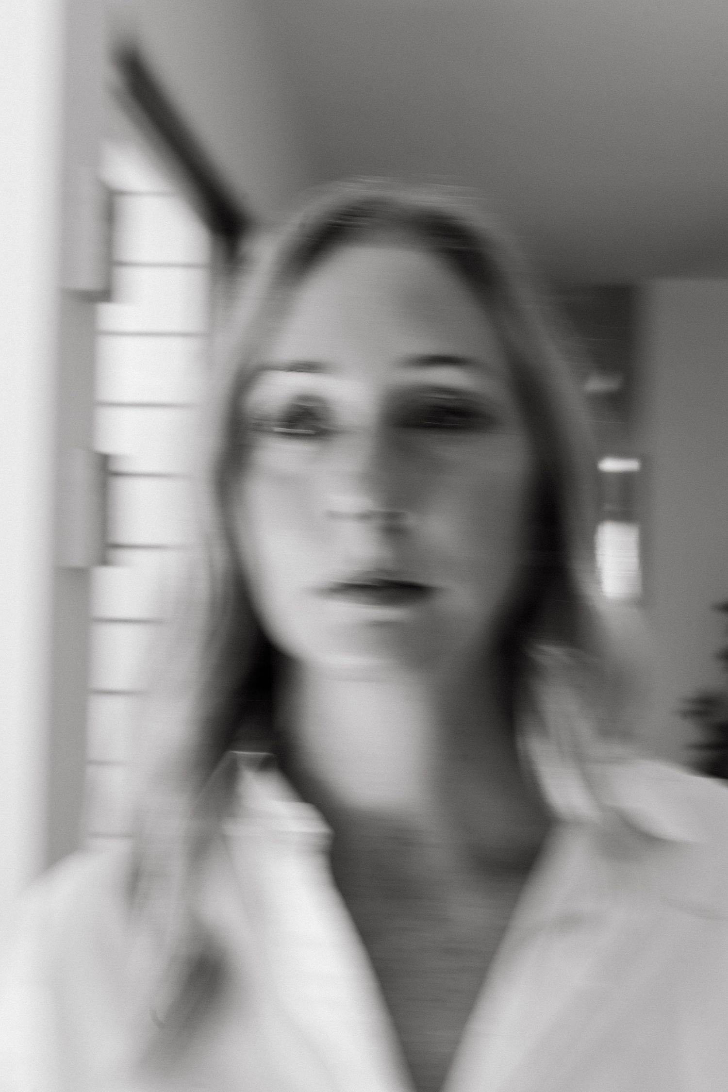 Blurry black and white portrait of a woman with long hair wearing a white shirt, standing in a hallway.